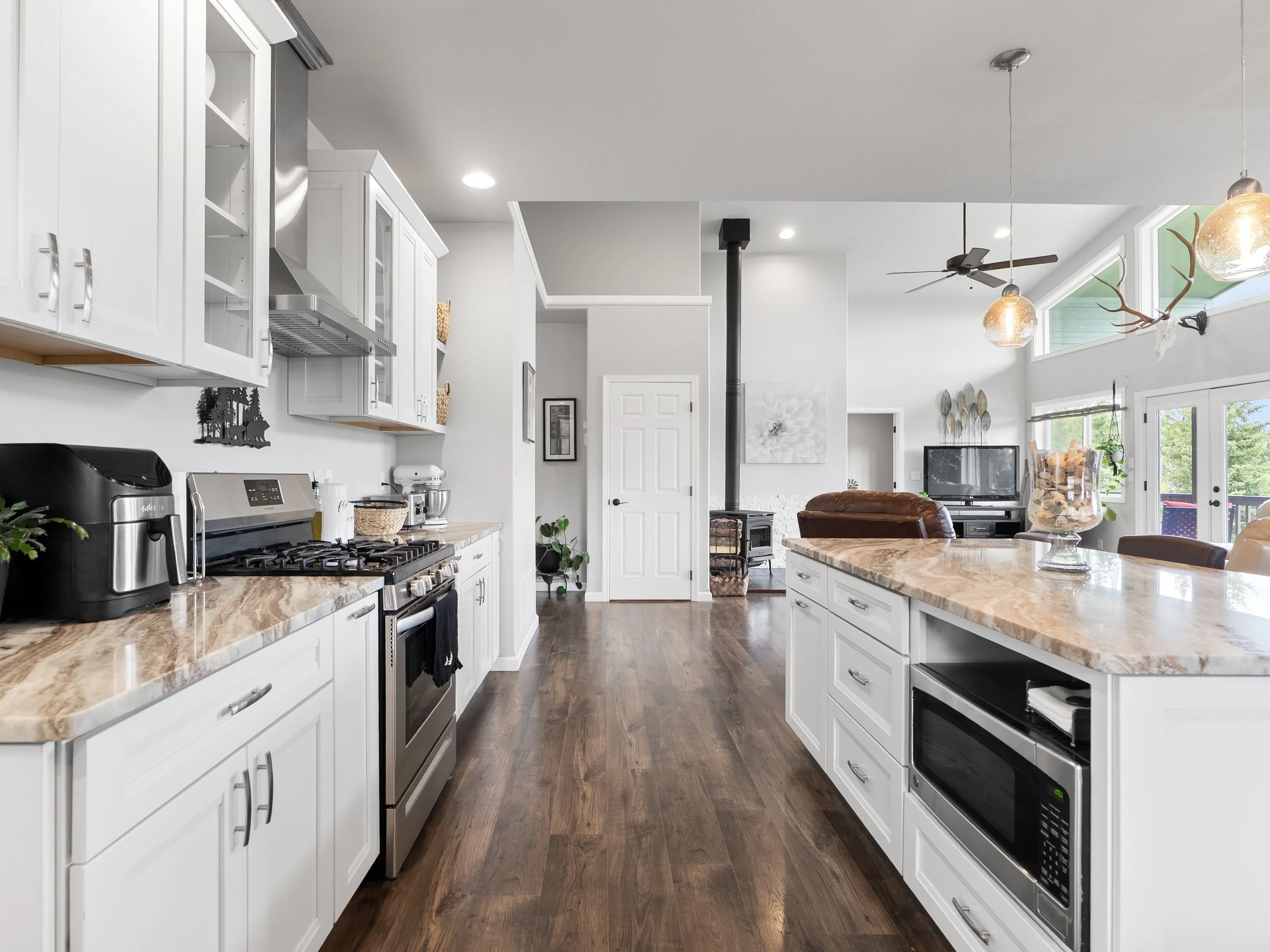 Modern kitchen with white cabinets, marble countertops, stainless steel appliances, dark hardwood floors, large windows, and a view of the living area with a stove, TV, and decorations.