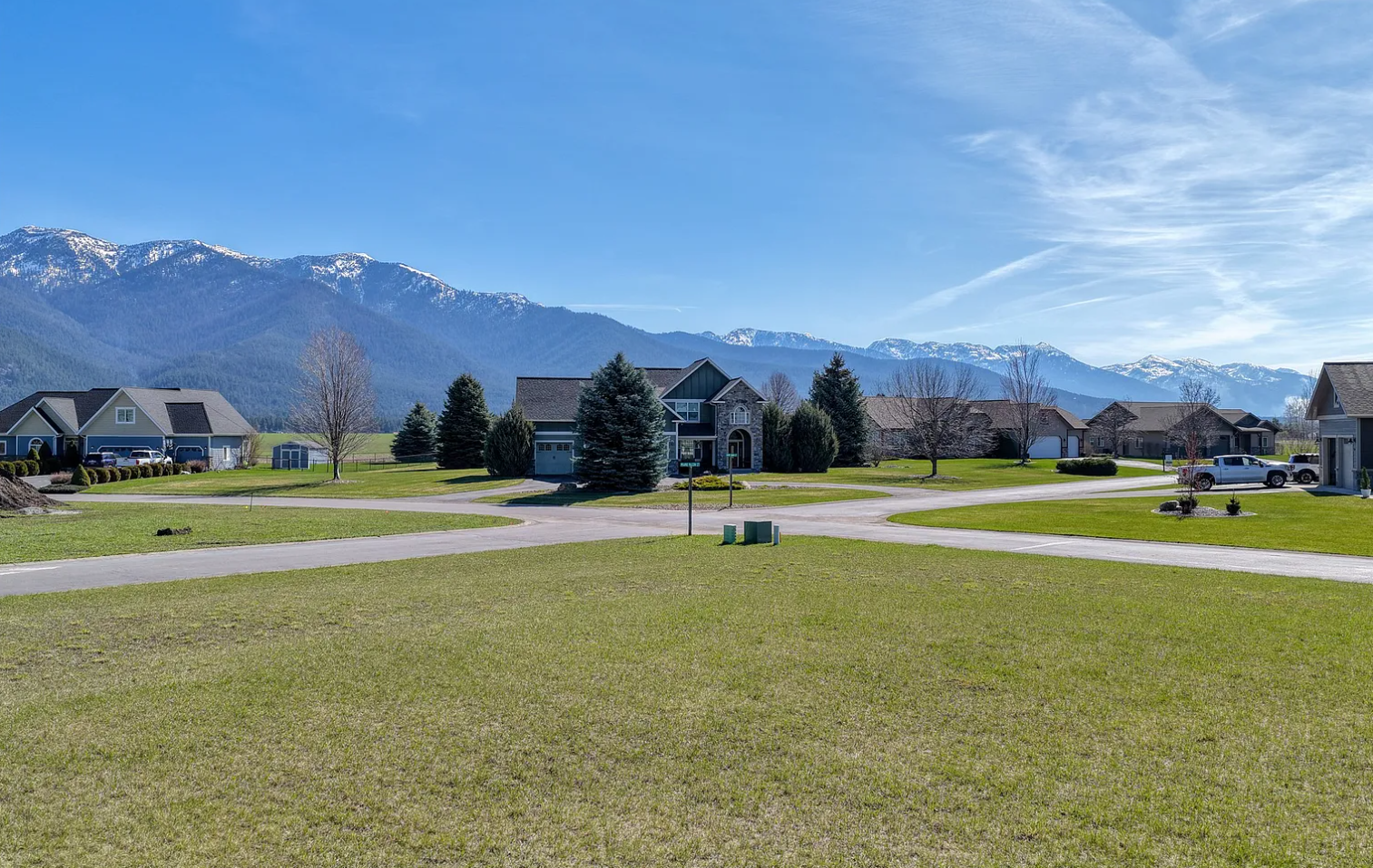 Suburban neighborhood with large houses, green lawns, and trees, set against snow-capped mountains under a clear blue sky.