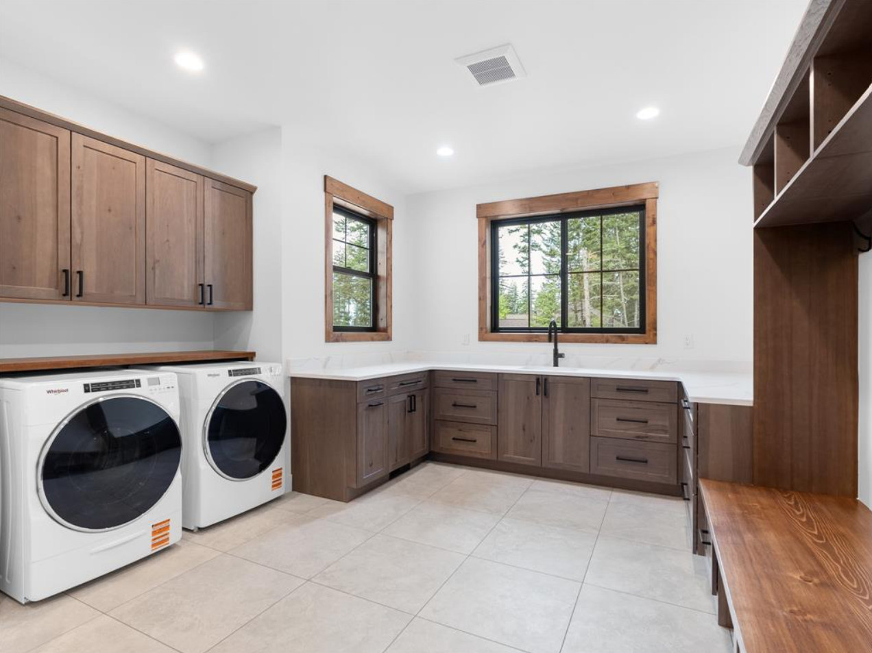 A modern laundry room featuring dark wood cabinetry, a white countertop, two large windows, a black faucet, and laundry appliances.