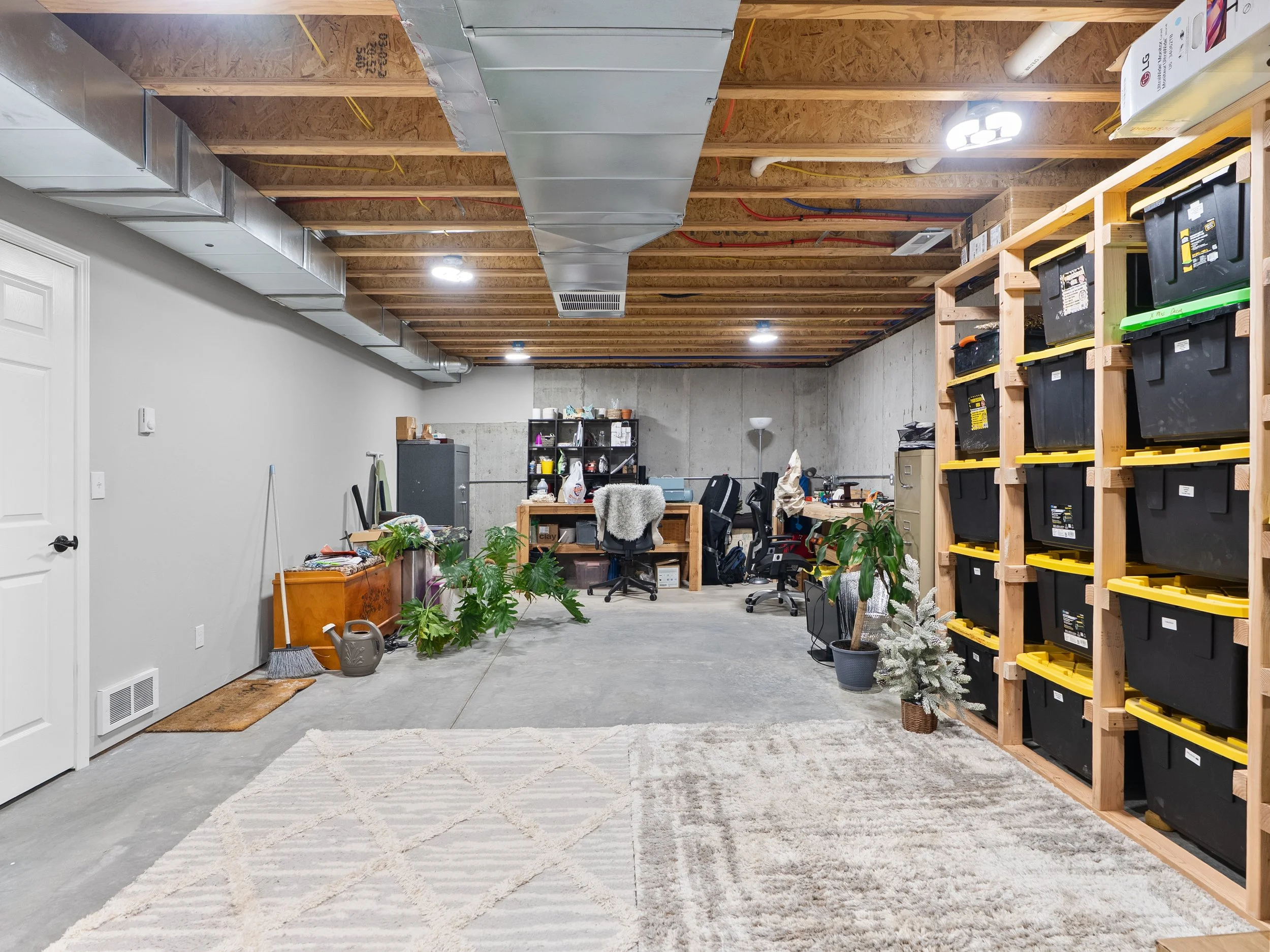 A partially finished indoor space with exposed wooden ceiling beams, ductwork, and electrical wiring. The left wall is white, and the floor is concrete with a light-colored rug. There's a black shelving unit with various items, a small table with pla
