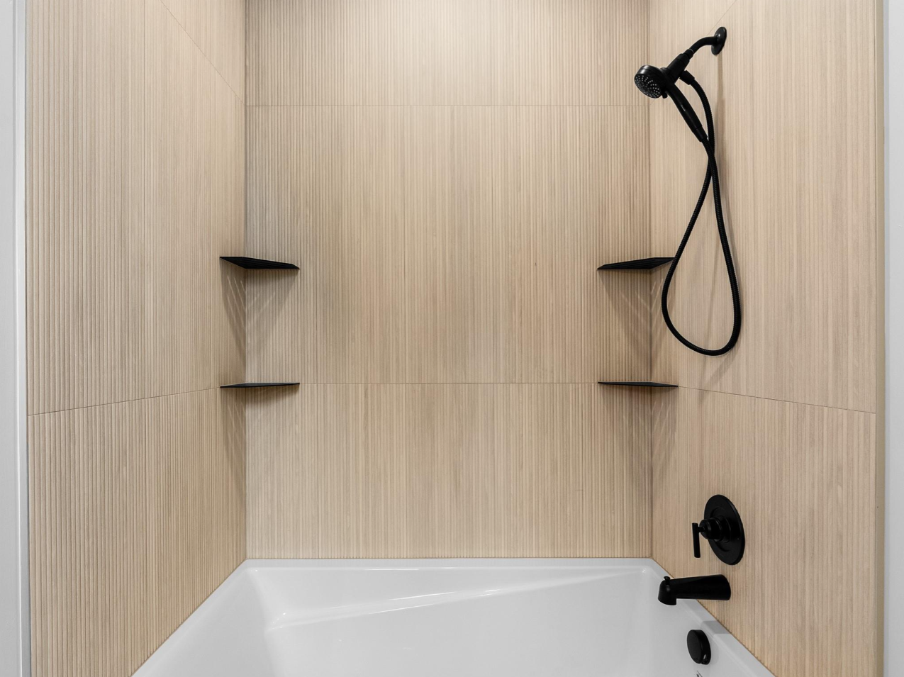 Modern shower with beige textured walls, black fixtures, handheld showerhead, and built-in corner shelves.