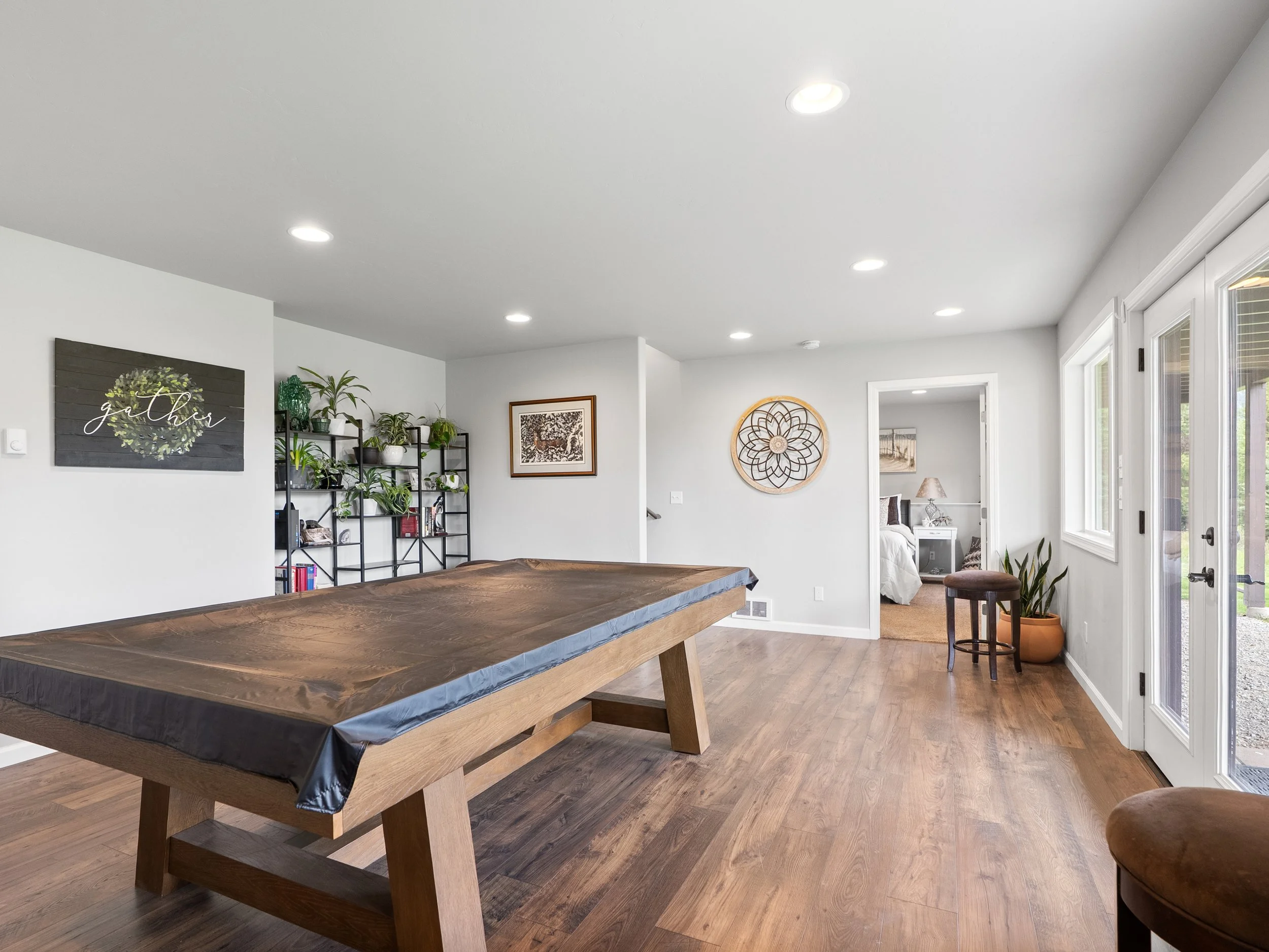 A living room with a large wooden table covered with black plastic, a wall decorated with framed art, bookshelf with plants, a window, and beige walls.