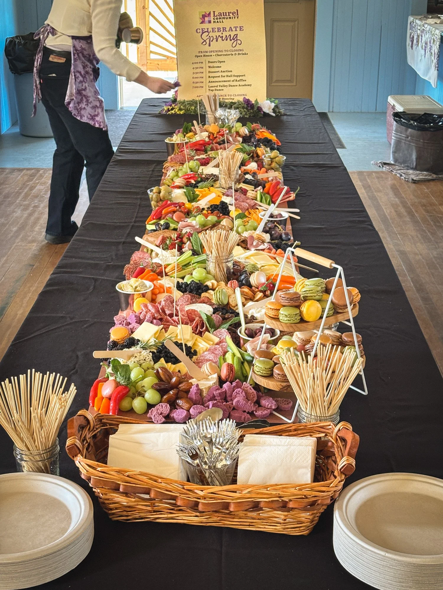 A table set up with a variety of colorful snacks including fruits, vegetables, cheese, meats, and crackers, with plates and napkins arranged in front, faux flower arrangements, and a yellow poster about a spring celebration at Laurel Community Hall in the background.