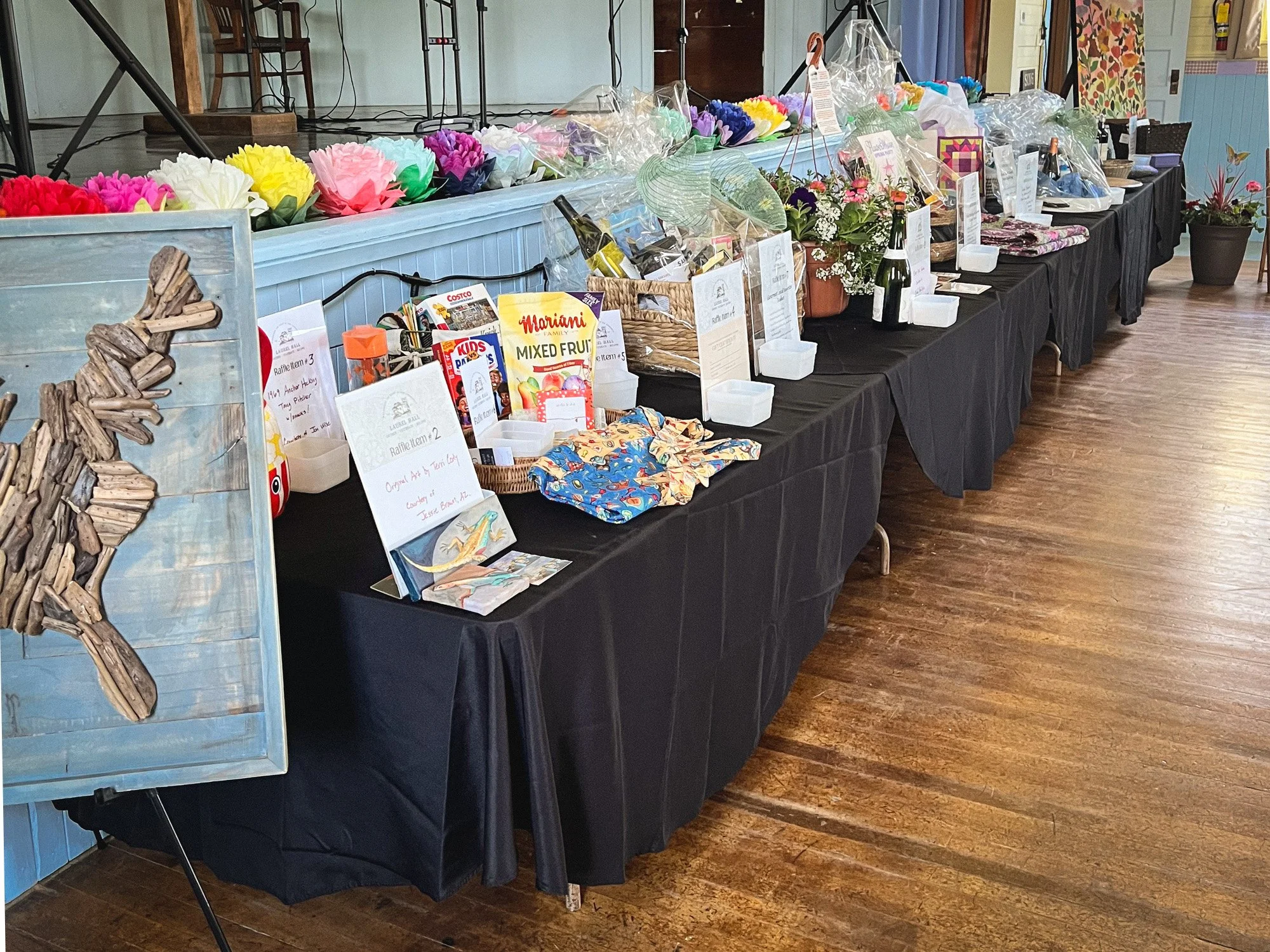 A long table covered with a black tablecloth displays various items for , including colorful paper flowers, gift baskets, bottles of wine, and informational signs. The table is inside a building with wooden floors and a light blue wall in the background.