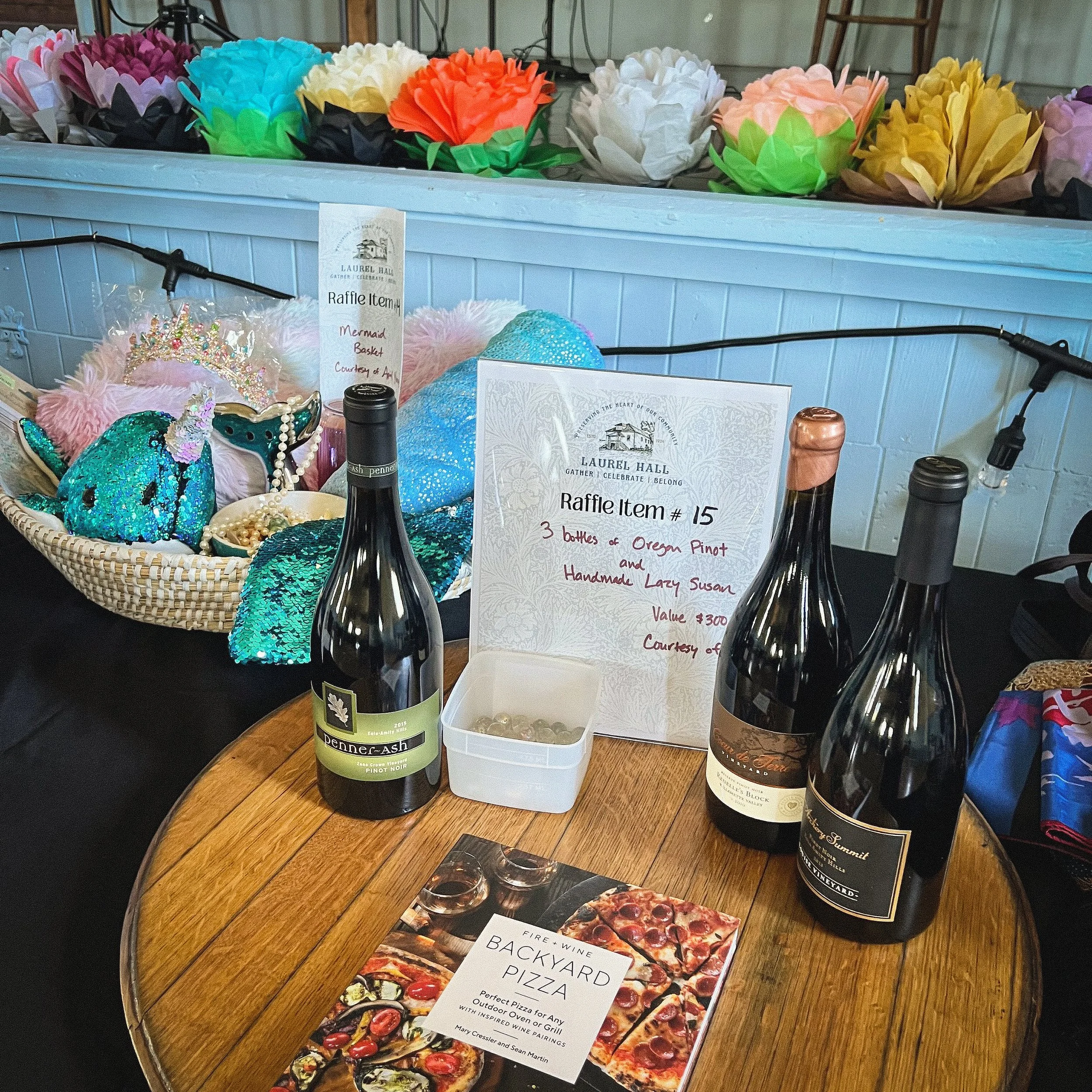 A table with a basket of colorful paper flowers, bottles of wine, a white container with raffle tickets, and a Backyard Pizza book.
