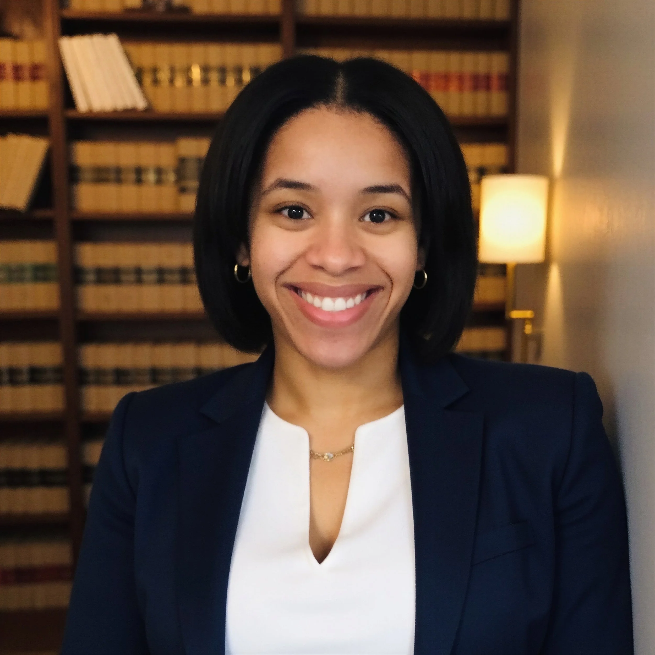 A woman with shoulder-length black hair smiling in a professional setting, standing in front of a bookshelf and a lit lamp.