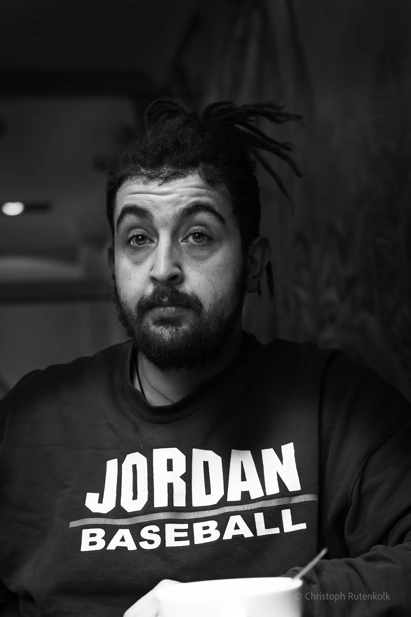 A man with dreadlocks and facial hair sitting at a desk, wearing a black sweatshirt with 'JORDAN BASEBALL' printed on it, looking at the camera with a serious expression.