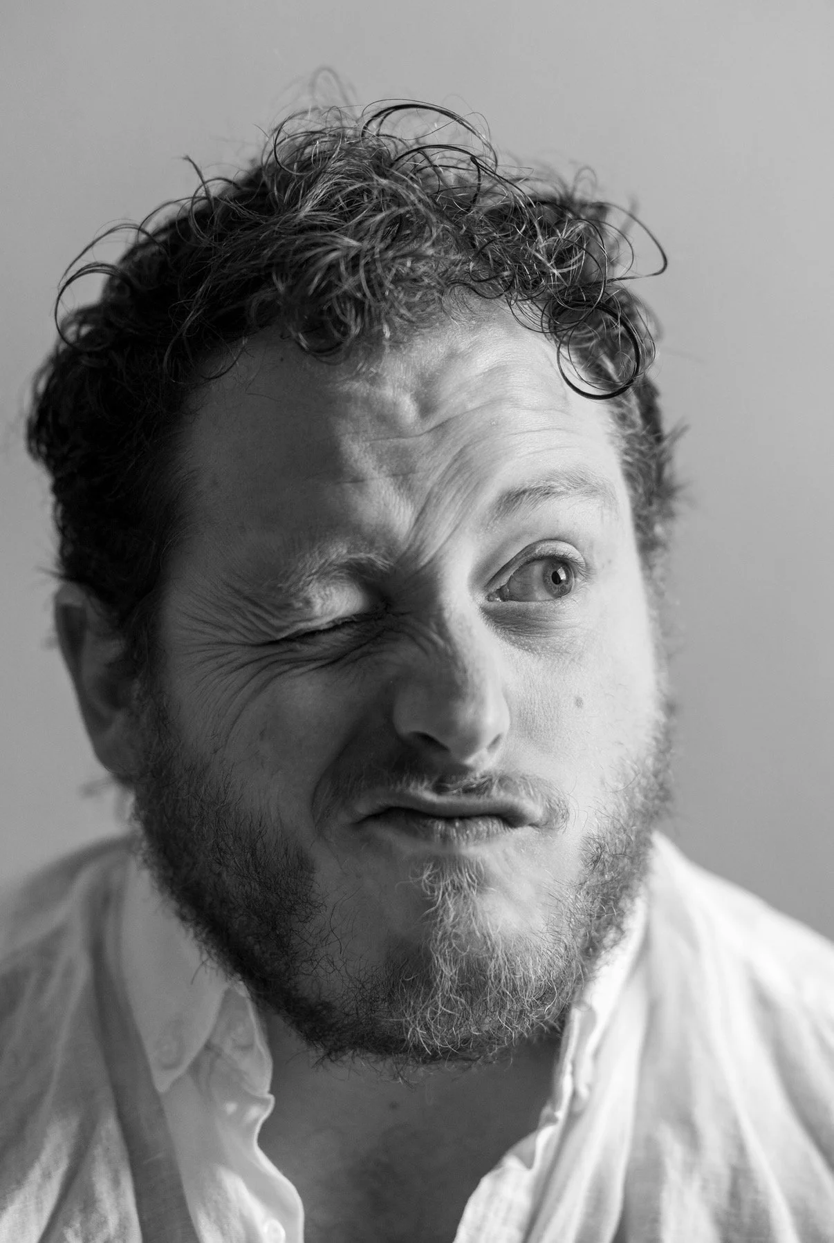 A black and white photo of a man with curly hair and a beard, making a playful facial expression with one eye squinted and his mouth slightly open.