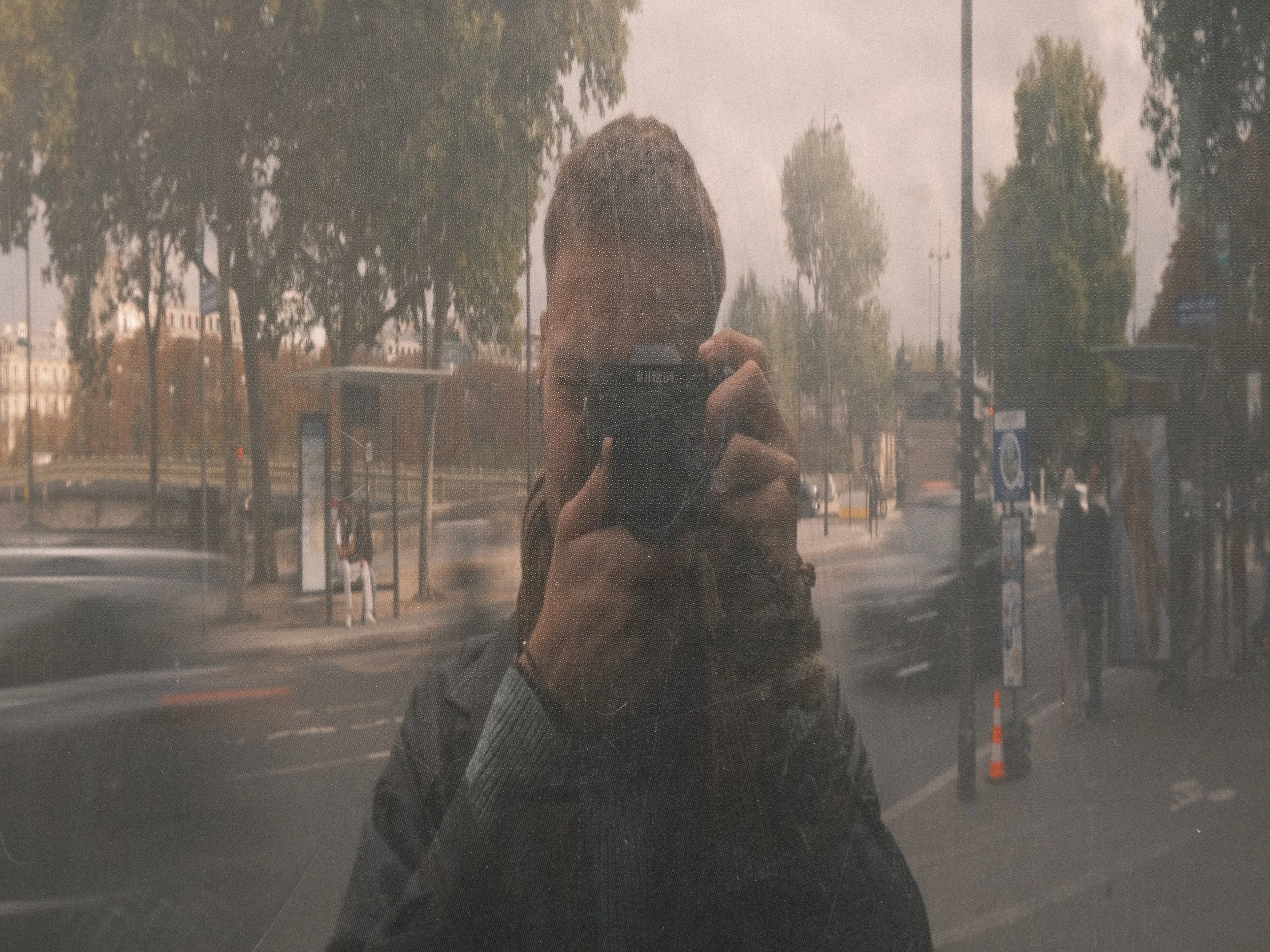 Person taking a photo of themselves in a mirror, with trees and buildings visible through a window behind them.