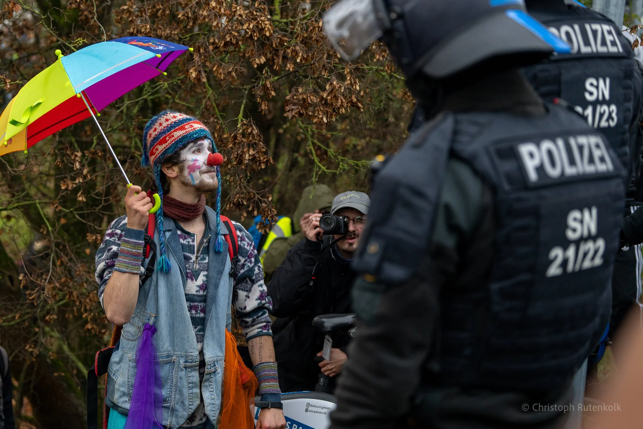 Clown with face paint, red nose, colorful hat, and rainbow-colored umbrella talking to a police officer in uniform and helmet outdoors, with trees and other people in the background.