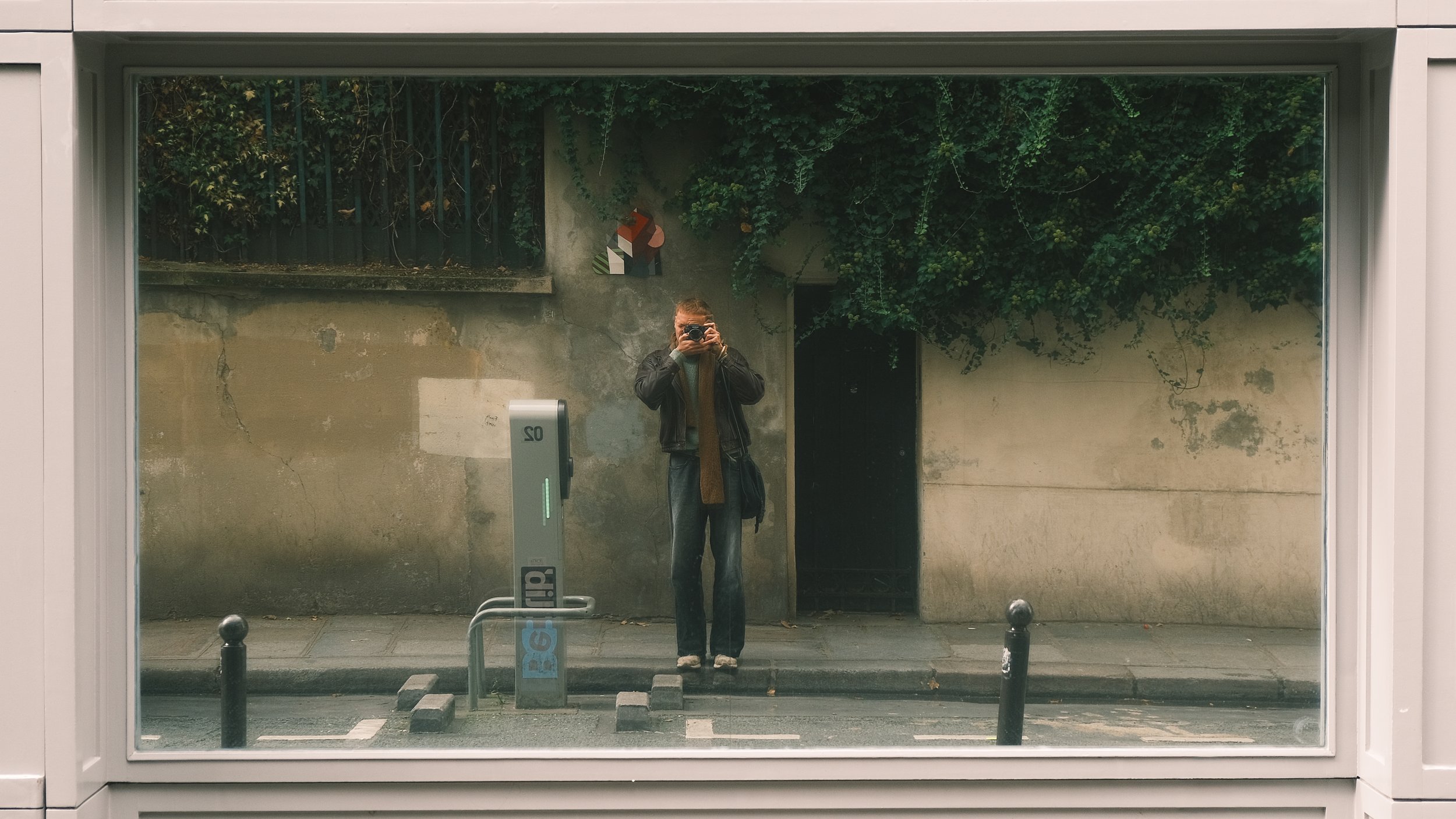 Reflection of a person taking a photo in a large street-side mirror, surrounded by greenery and a gray wall.