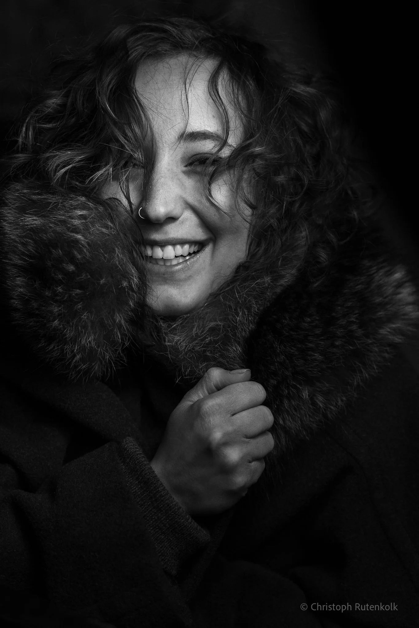 Black and white photo of a woman smiling with curly hair, wearing a fur-lined coat, hand gripping the coat's collar.