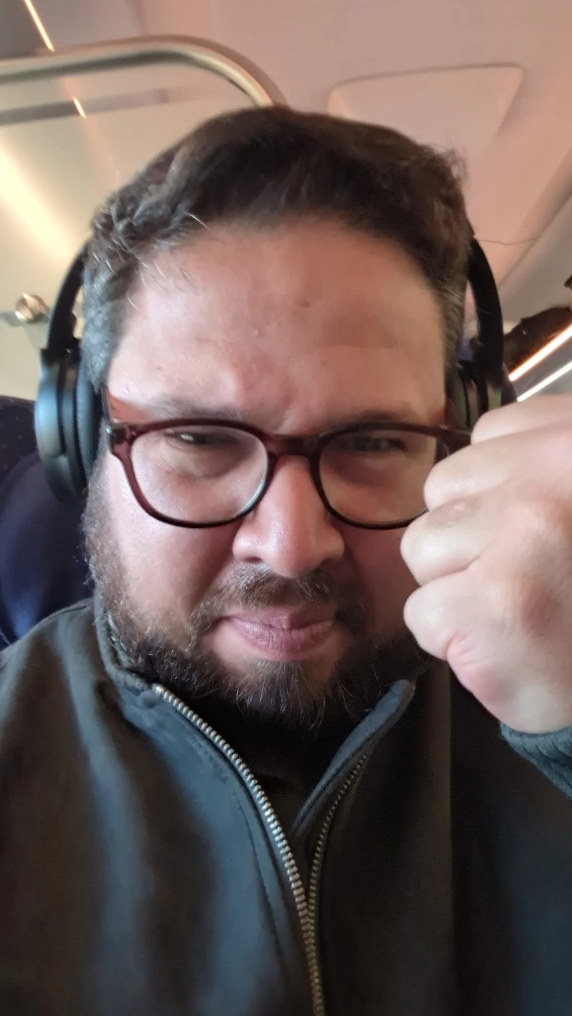 A man with dark hair, glasses, and a beard is wearing headphones and making a fist with his right hand. He is wearing a gray jacket and is sitting in what appears to be an airplane.
