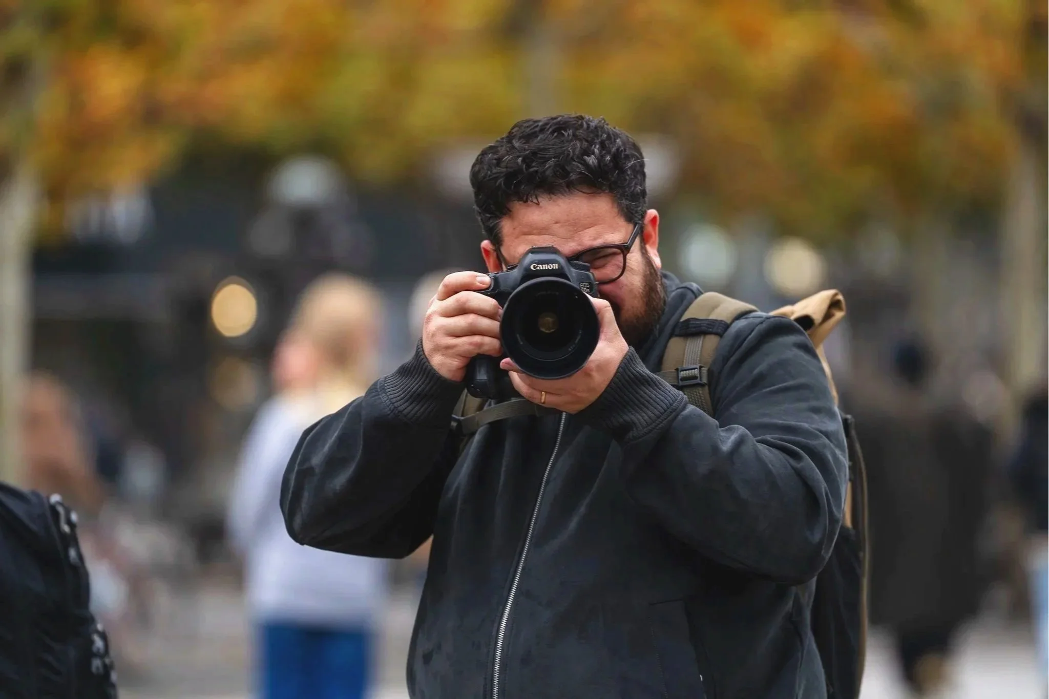 Jose Villacreses is taking a photo with a Canon camera outdoors during fall. He is wearing a black jacket with a backpack, and there are blurred autumn-colored trees and people in the background. Photo Christoph Rutenkolk