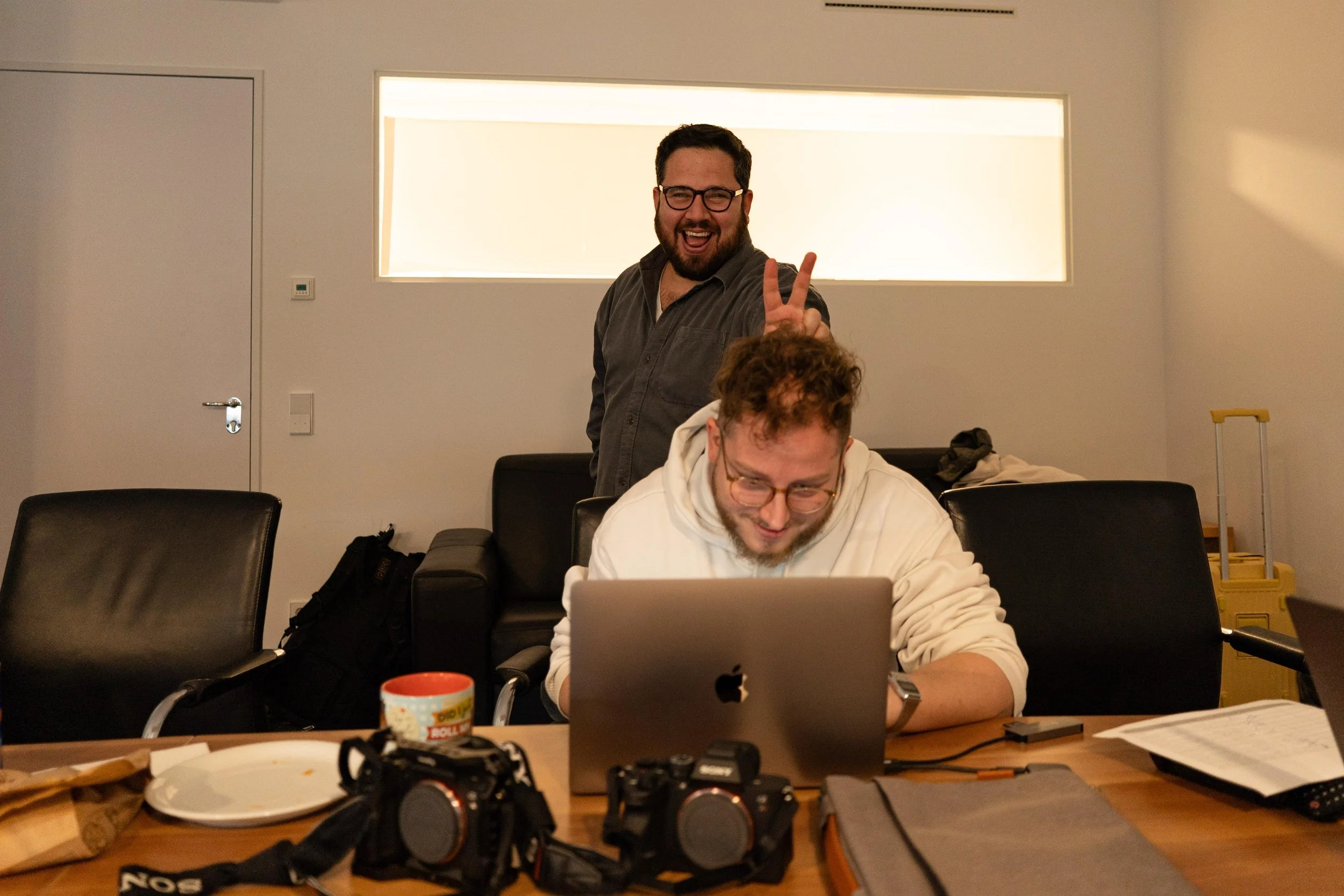 Two men working at a table in an office, one standing and making a peace sign, the other sitting and working on a laptop.