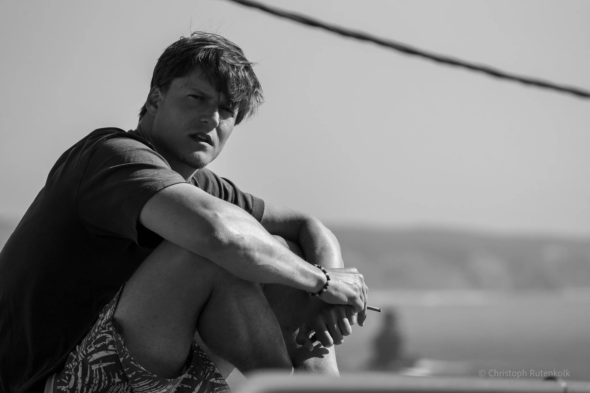 A black and white photograph of a young man sitting outdoors, looking pensively into the distance, with a blurred horizon background.