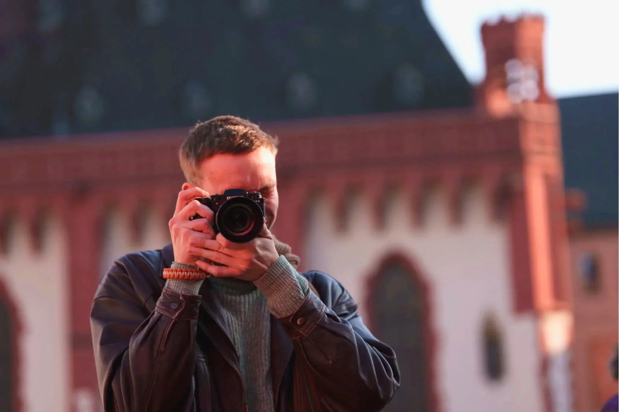 A person with short hair, wearing a layered jacket and sweater, holds a camera up to take a picture, with a blurred building with arches in the background.