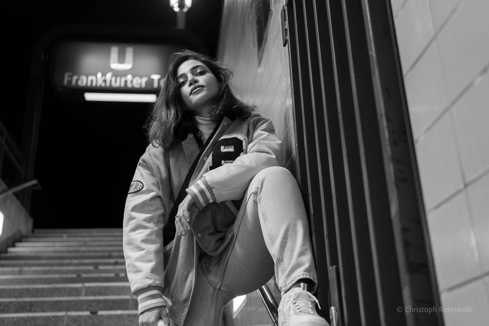 A young woman with wavy hair sitting on stairs at a subway station near a metal railing. She is wearing a varsity jacket, light-colored pants, and sneakers, looking directly at the camera with a serious expression. There is an illuminated sign in the background that reads "Frankfurter T".