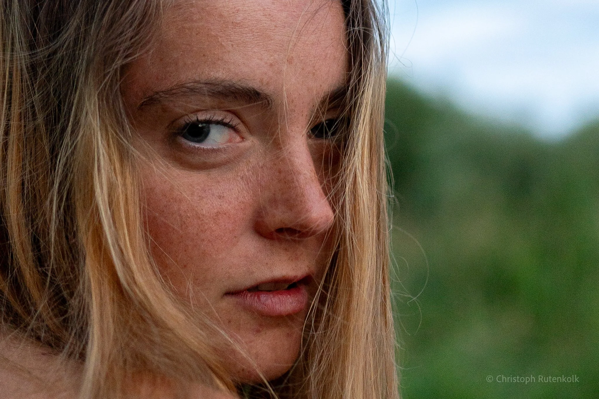 Close-up of a woman with blonde hair, blue eyes, and freckles, outdoors with a blurry green background.