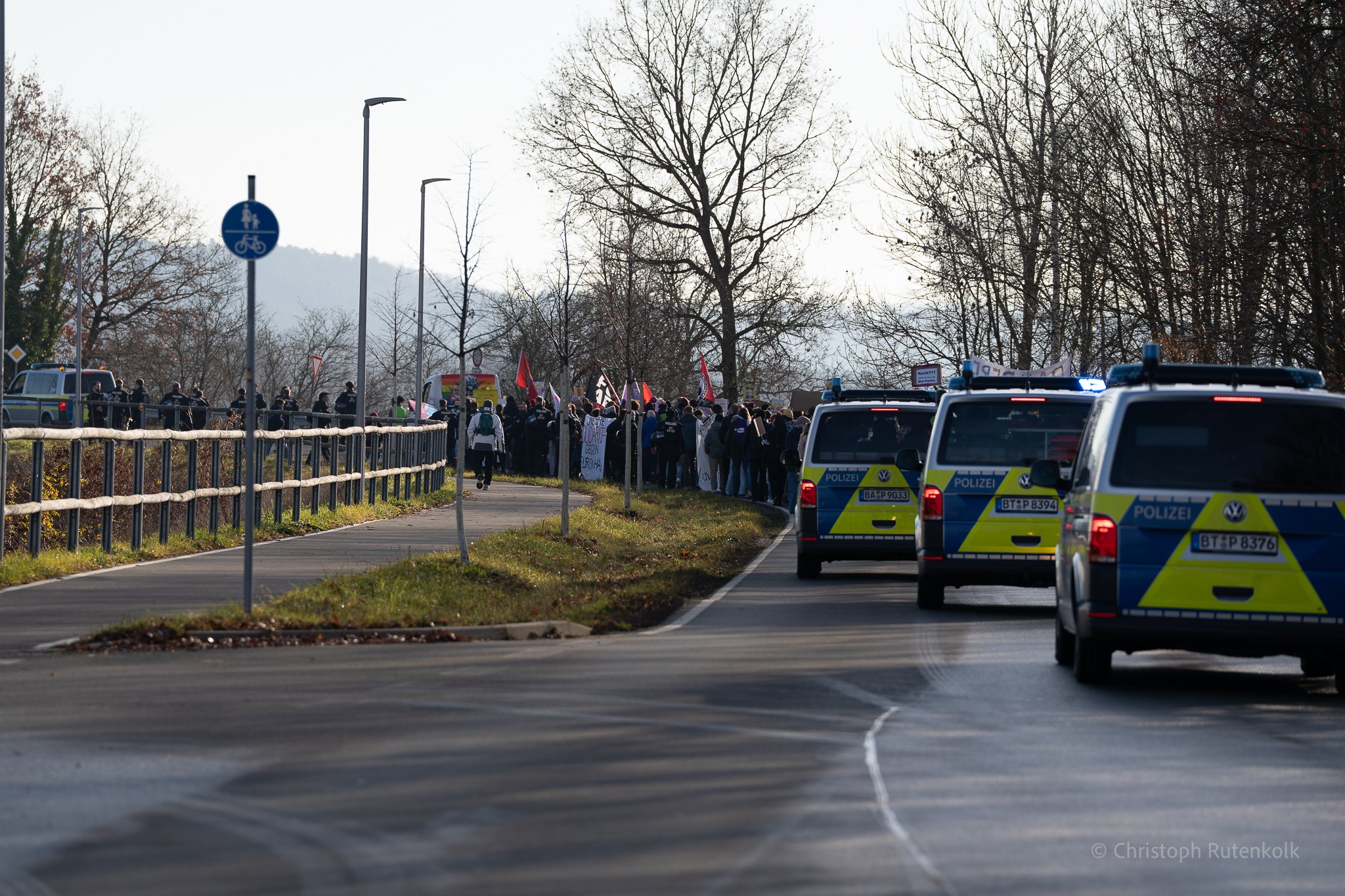 A group of protesters holding signs and flags walking on a sidewalk beside a police vehicle. Several police cars are parked on the road nearby. The scene takes place during daytime with leafless trees in the background.