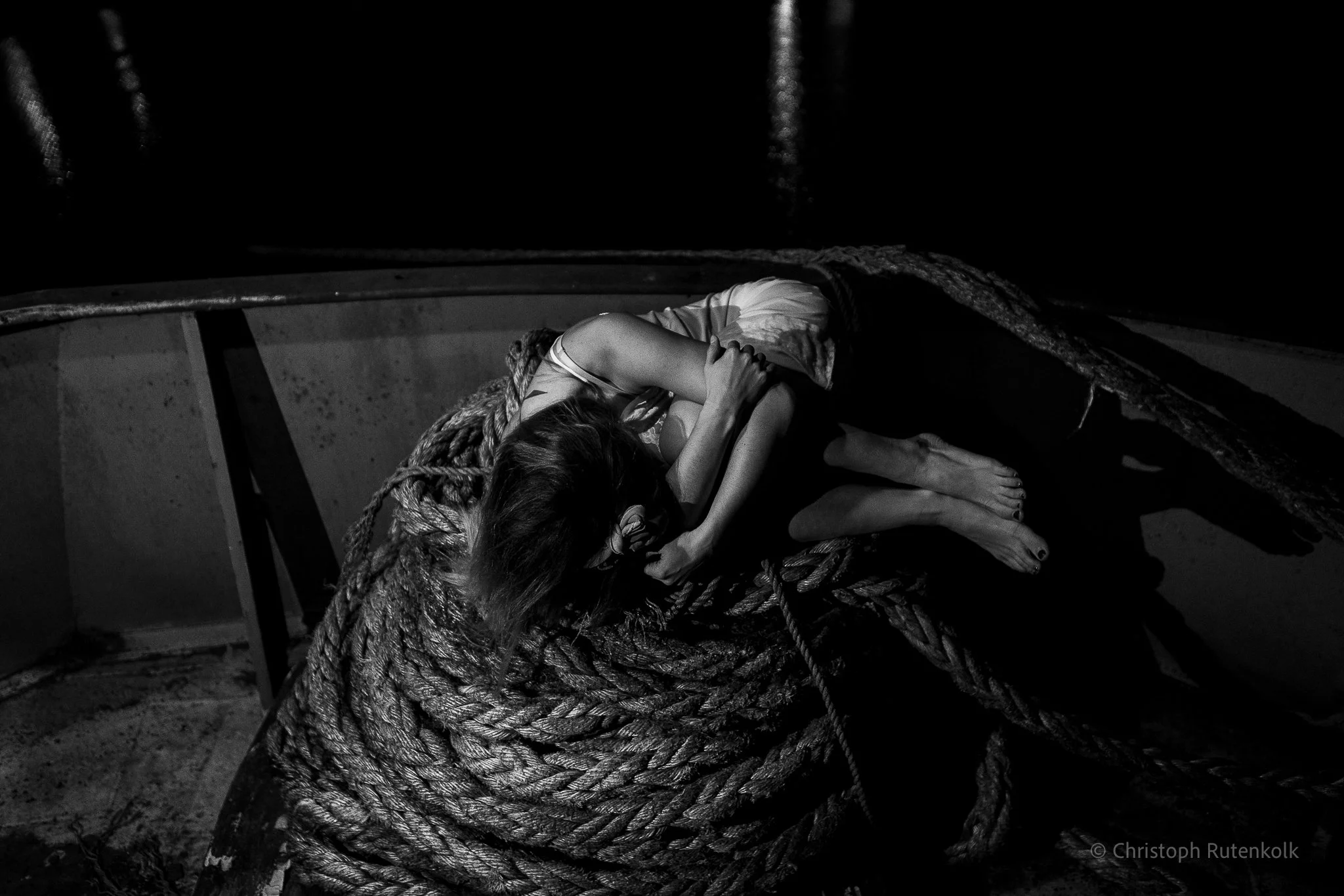 A person curled up on a boat's deck, hugging their knees, with long hair covering their face, surrounded by thick ropes in a dark environment.