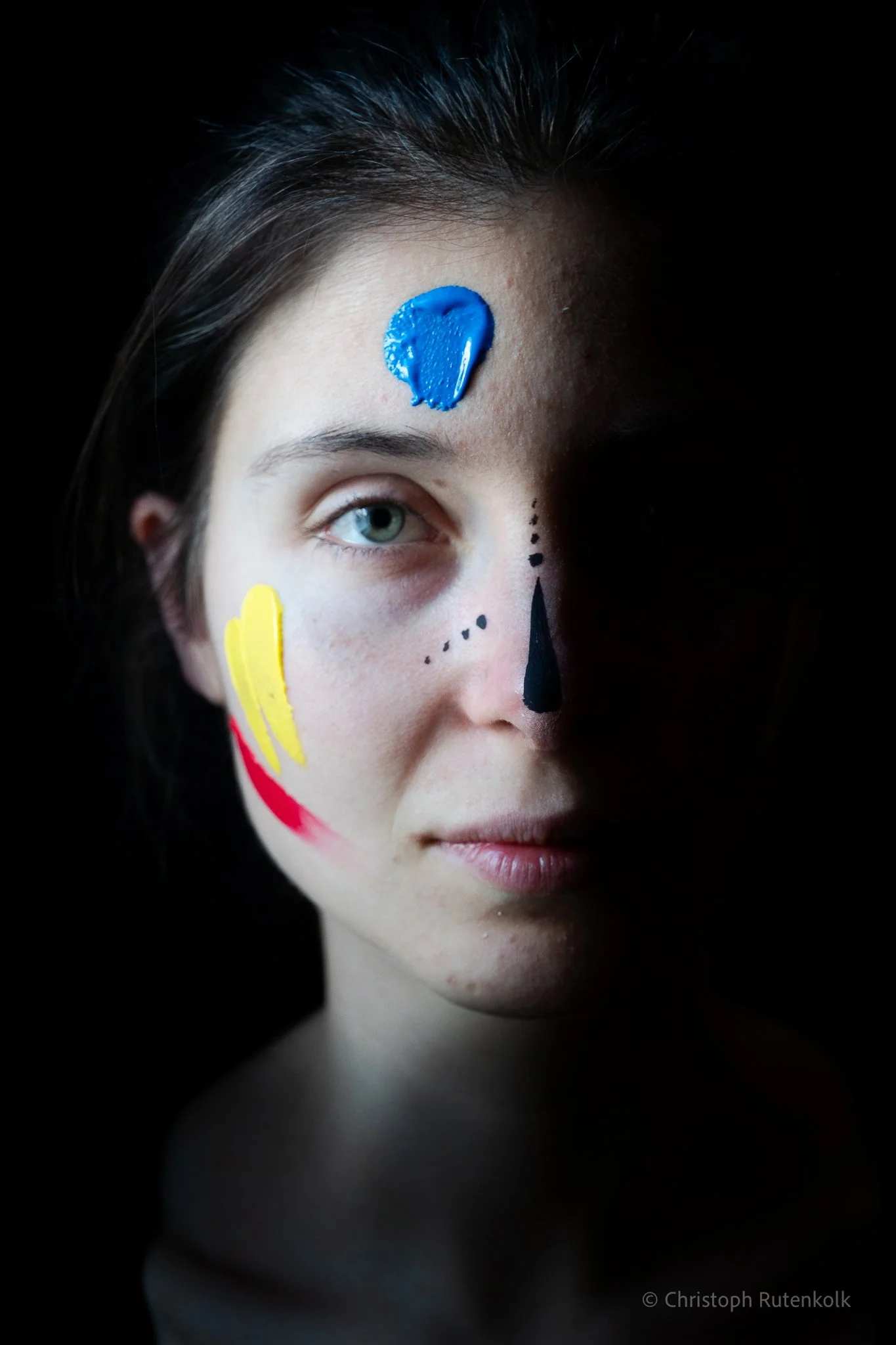 A woman with face paint resembling a sugar skull, with a blue heart-shaped paint on her forehead, black teardrop and dotted designs on her face, and colorful paint on her cheeks, set against a dark background.