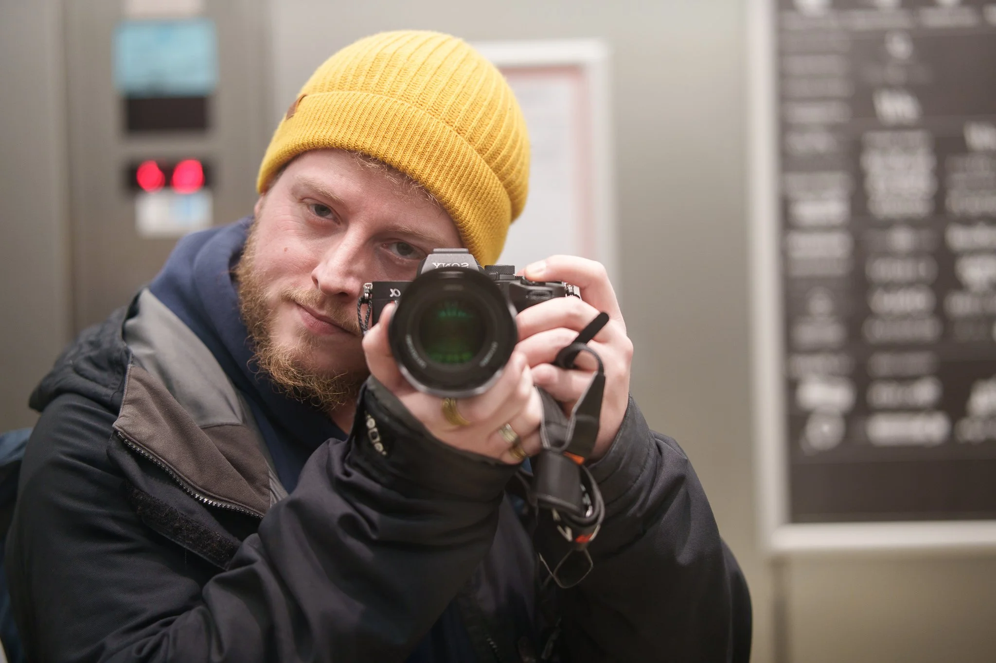 A man wearing a yellow knit cap taking a selfie with a camera in an indoor setting.