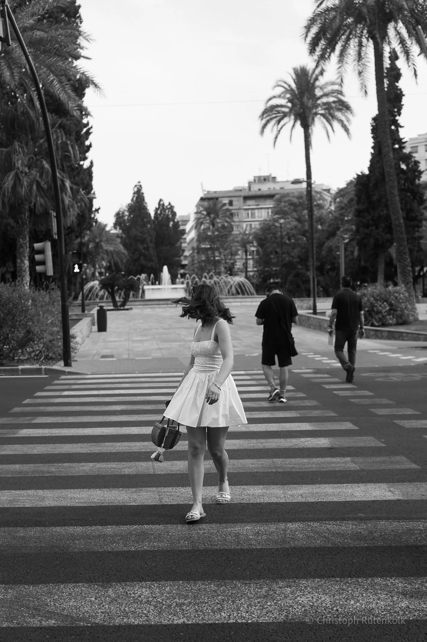 A woman in a white dress crossing a pedestrian crosswalk in an urban park area with palm trees, fountains, and buildings in the background, captured in black and white.
