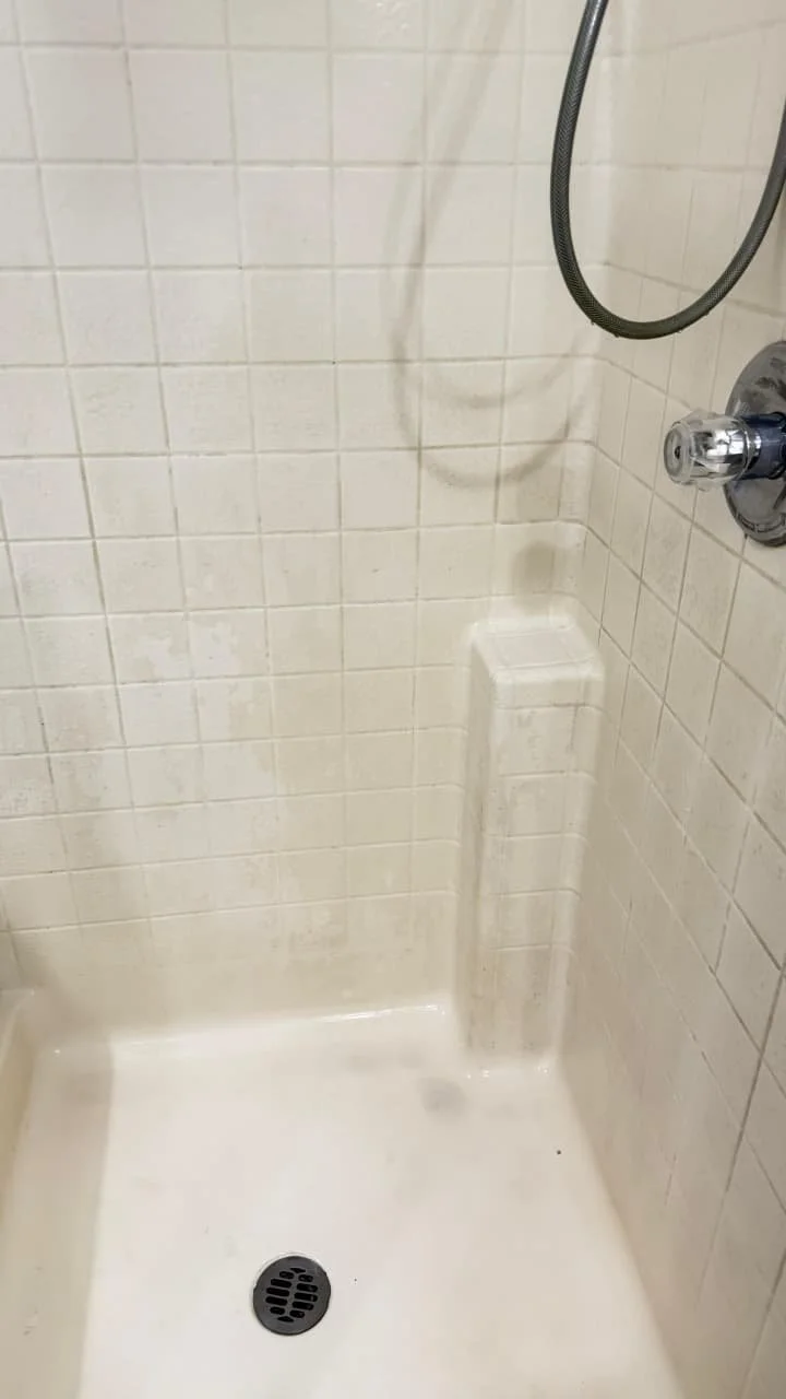 Empty shower stall with cream-colored tiled walls, a drain at the bottom, and a mounted showerhead with a flexible hose.