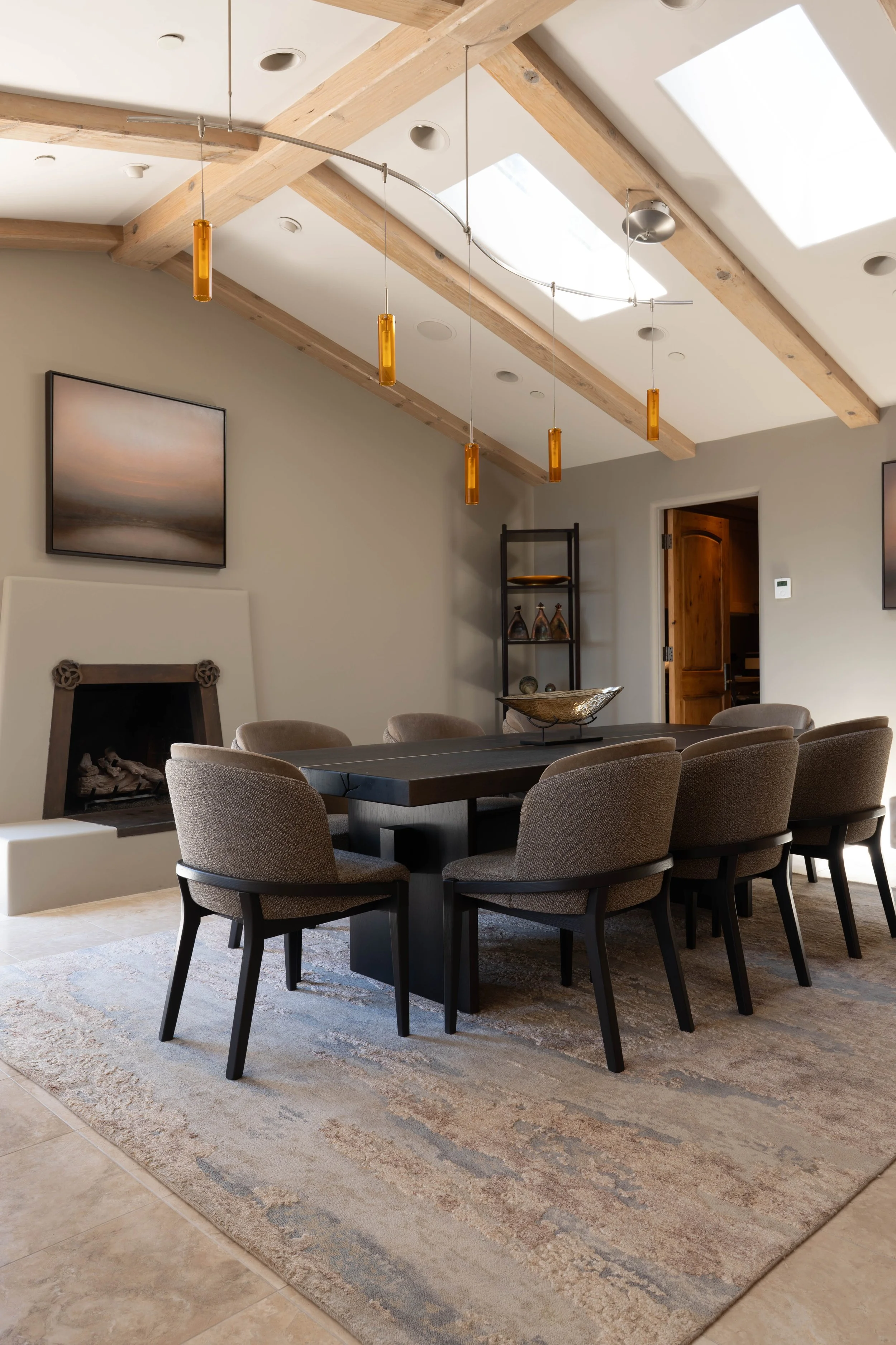 A dining room with a large black table surrounded by eight upholstered beige chairs. A decorative bowl is at the center of the table. Gray walls feature a flat-screen TV and a small black shelf with decorative items. The room has a fireplace and artwork. The ceiling has large wooden beams and skylights, with modern hanging light fixtures.