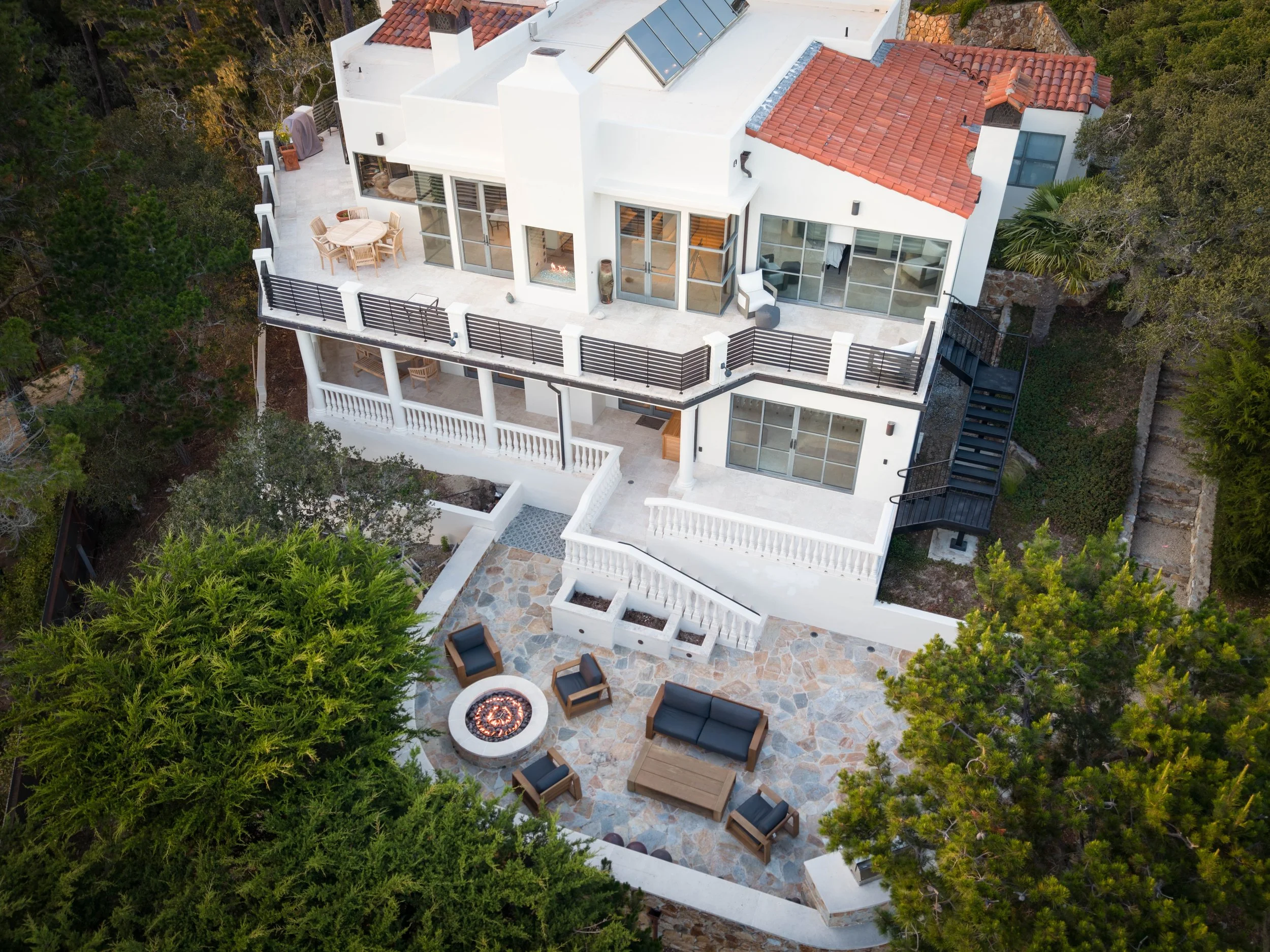 An aerial view of a modern white house with terracotta roof tiles, a rooftop solar panel array, and multiple large glass windows and doors. The house features outdoor living spaces with seating, a fire pit, and is surrounded by lush green trees.