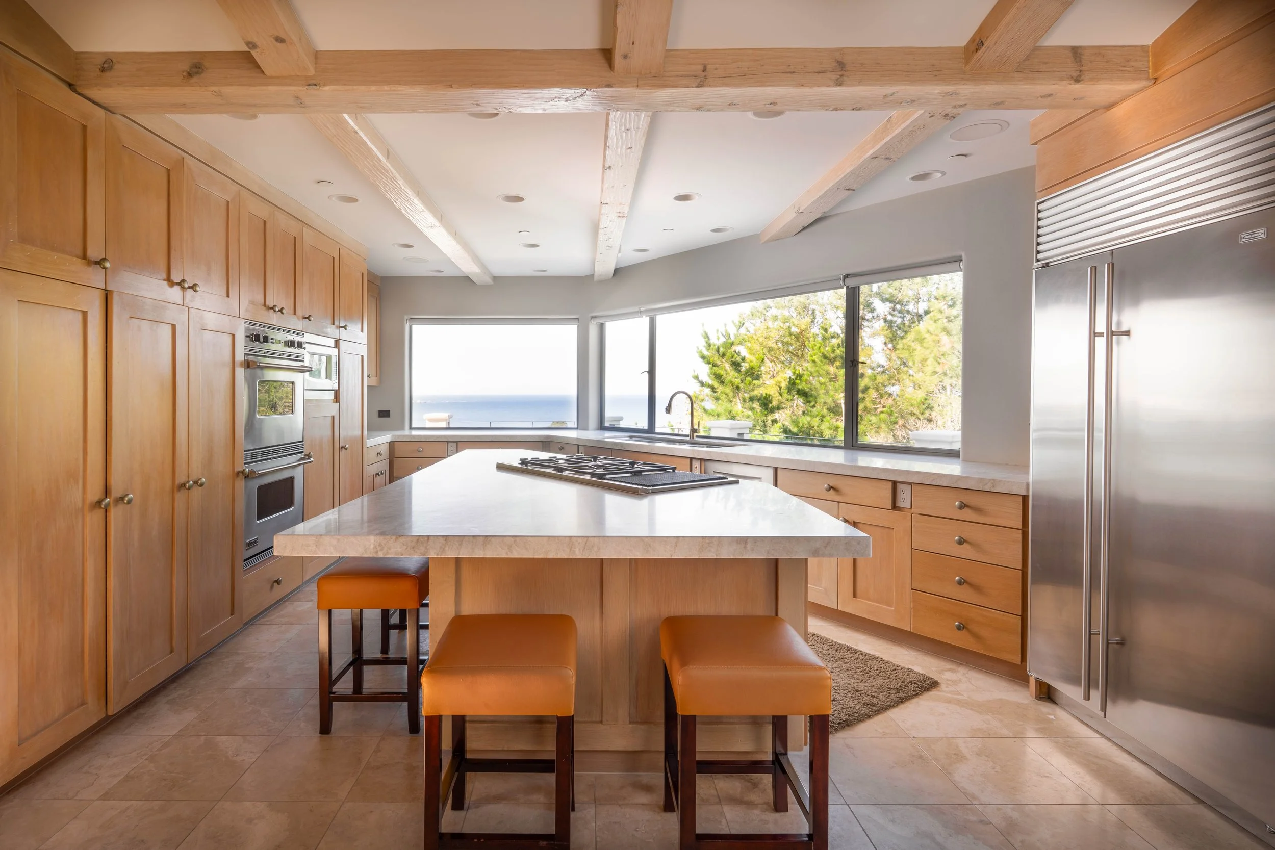 Modern kitchen with wooden cabinets, marble countertop island, stainless steel appliances, large windows showing outdoor greenery and ocean view, wooden ceiling beams, tan tiled floor, and three brown stools at the island.