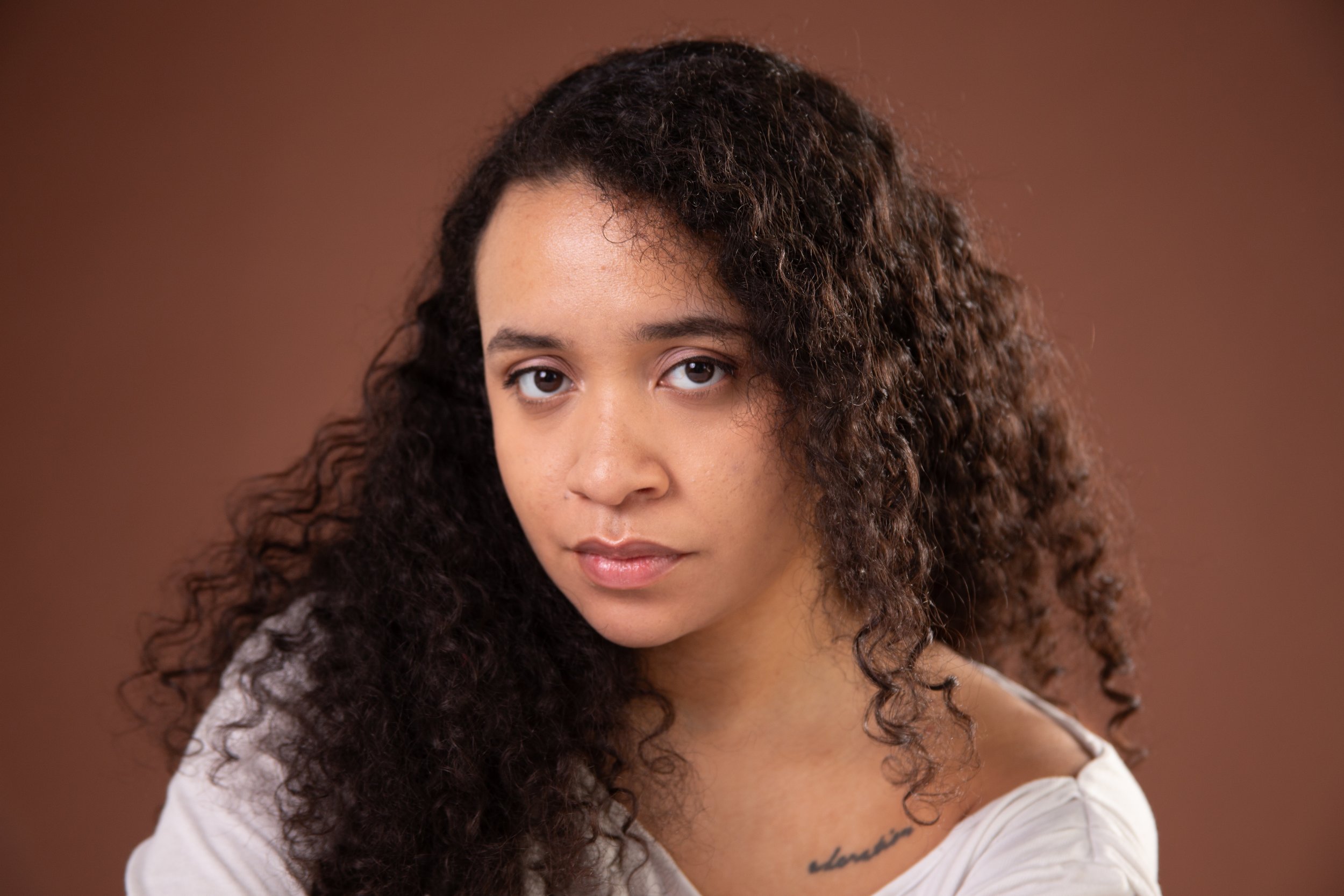 A young woman with curly dark hair and a neutral expression, wearing a white top, against a brown background.
