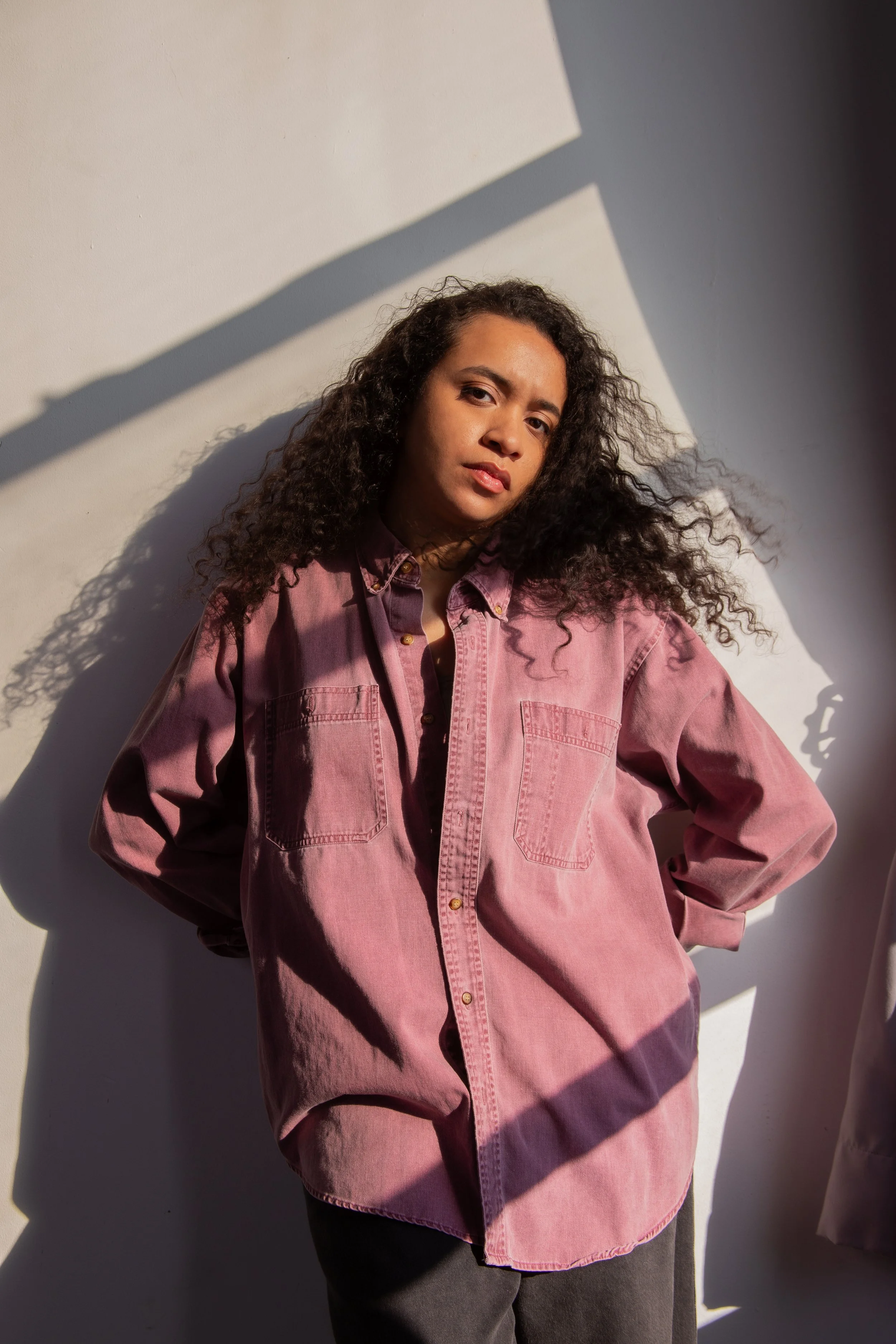 A woman with curly hair wearing a pink shirt standing against a white wall with shadows from the sunlight.