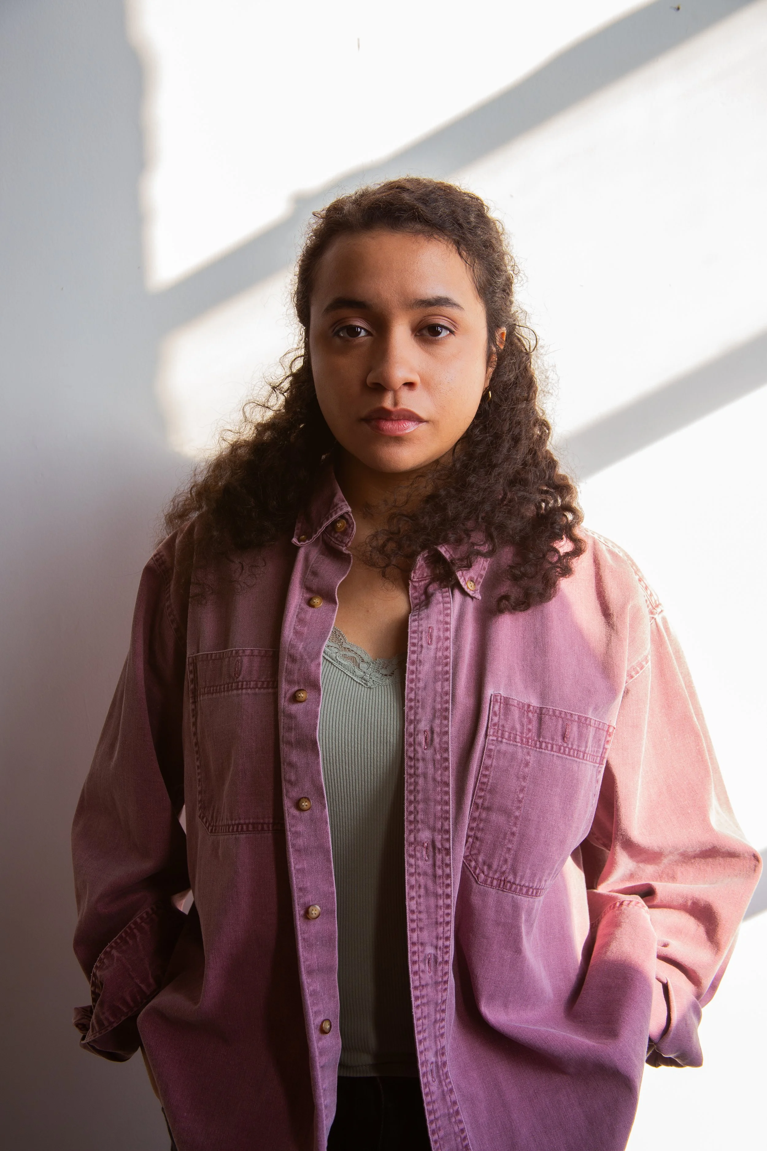 A woman standing against a white wall with shadow lines, wearing a light-colored tank top and an oversized pink button-up shirt with hands in pockets.