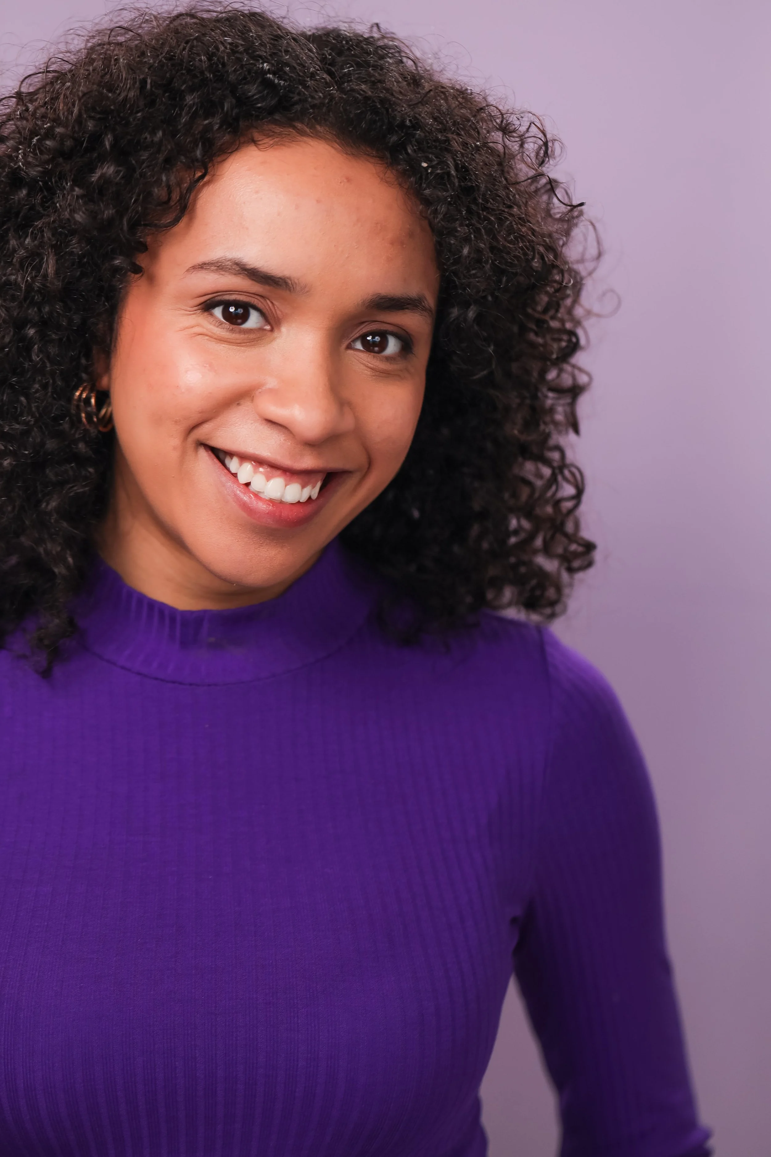A young woman with curly hair wearing a purple ribbed top, smiling against a light purple background.