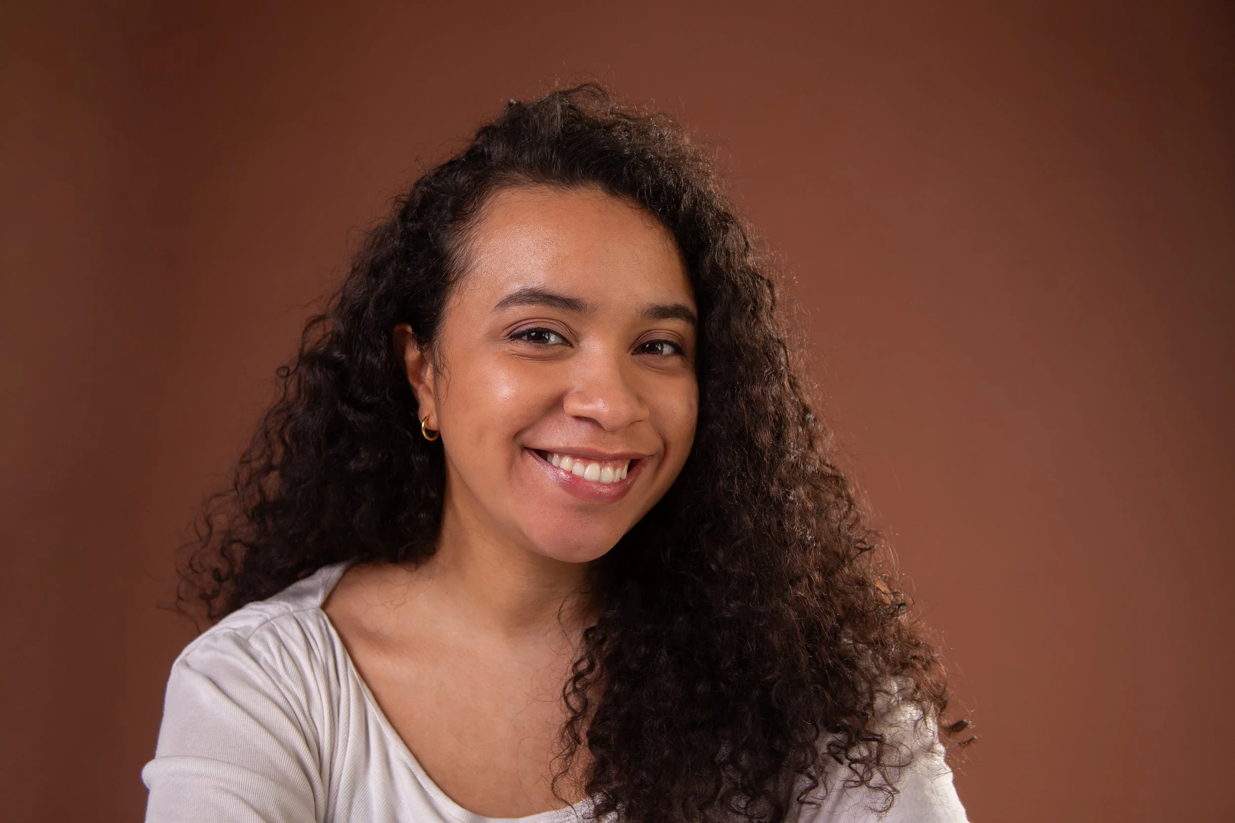 A smiling woman with curly dark hair, wearing a beige top, against a brown background.
