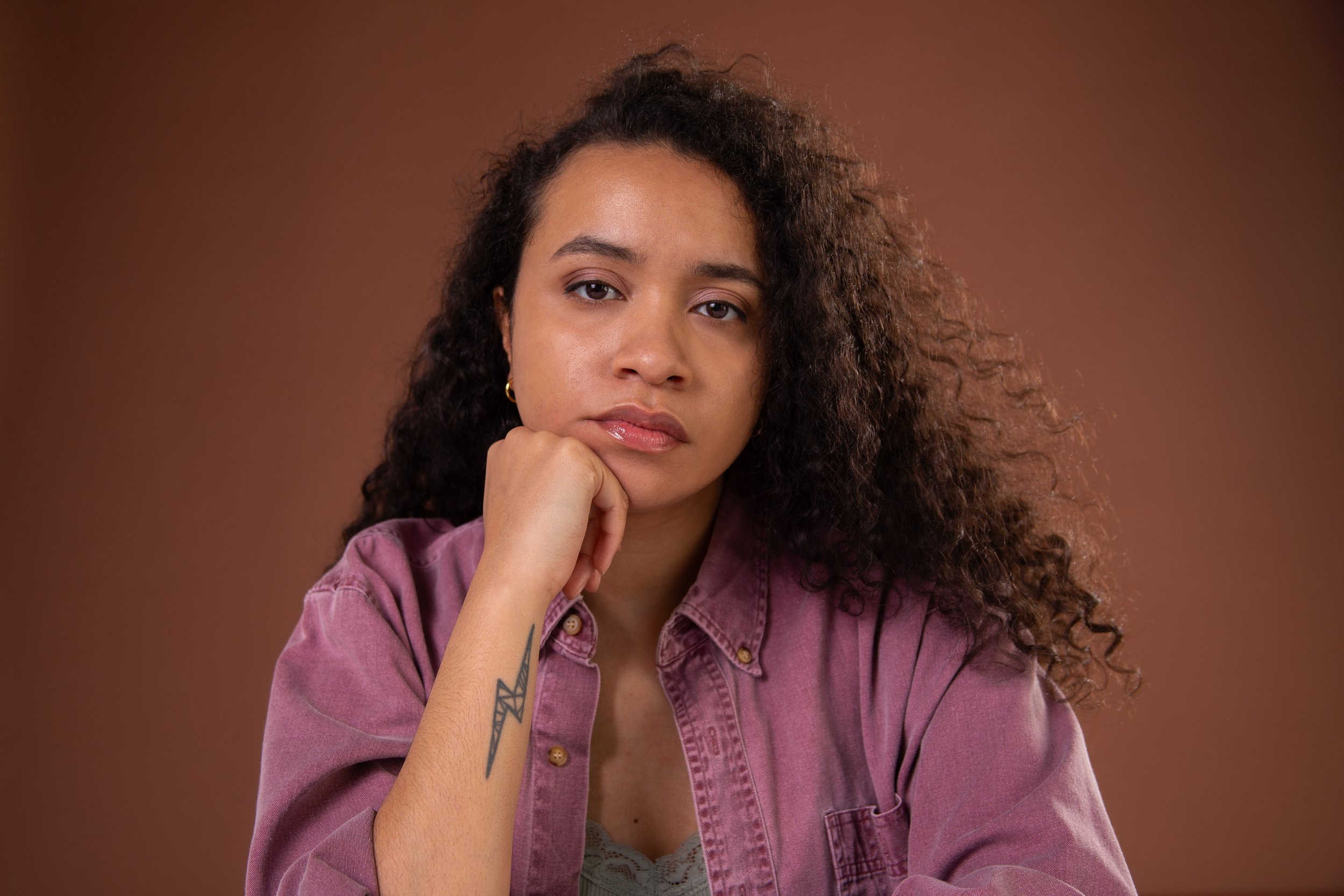 A woman with curly hair resting her chin on her hand, wearing a pinkish denim jacket, with a neutral expression against a brown background.