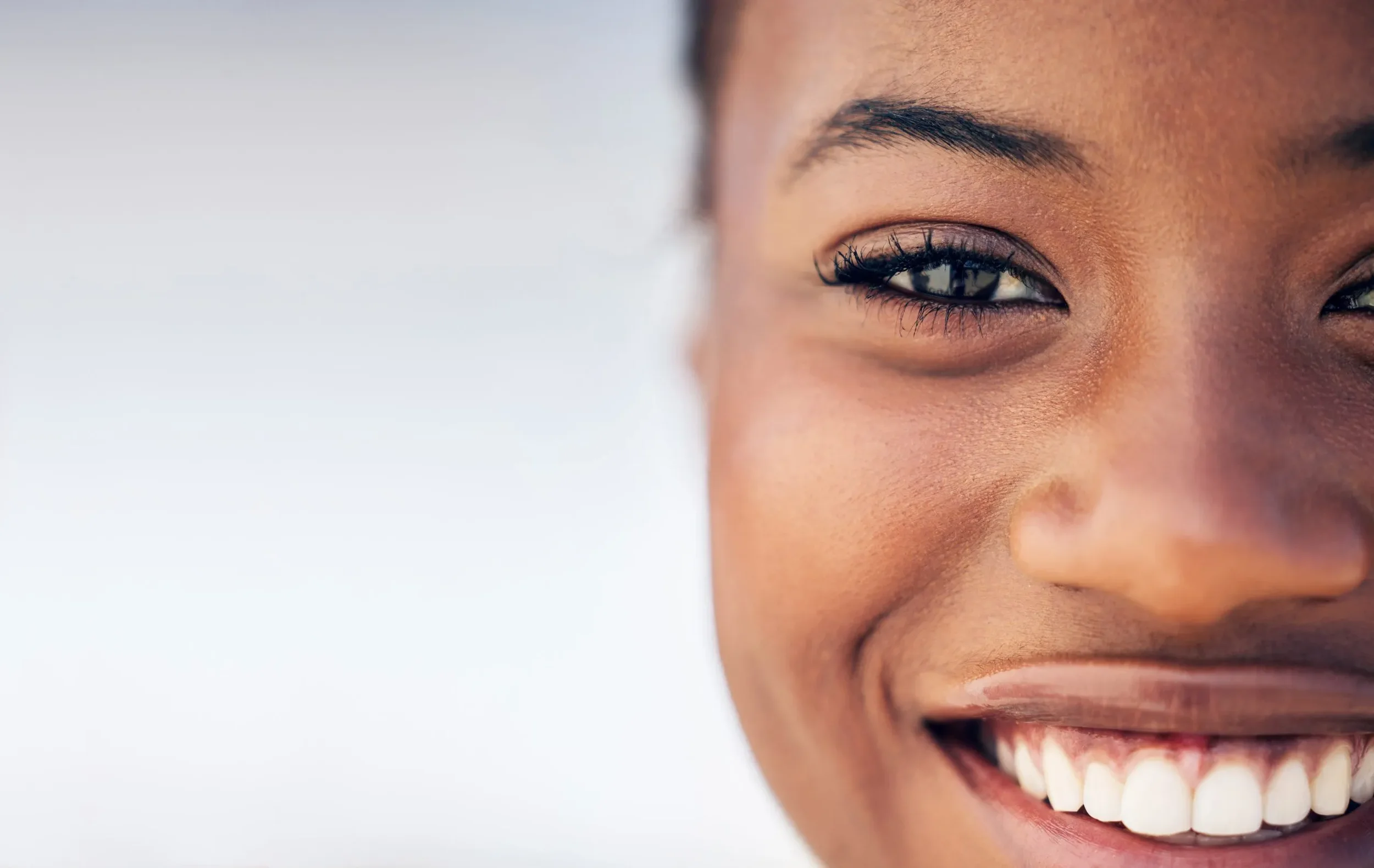 Close-up of a smiling young woman with dark skin and the right side of her face visible, showing her eye, nose, and mouth with white teeth.