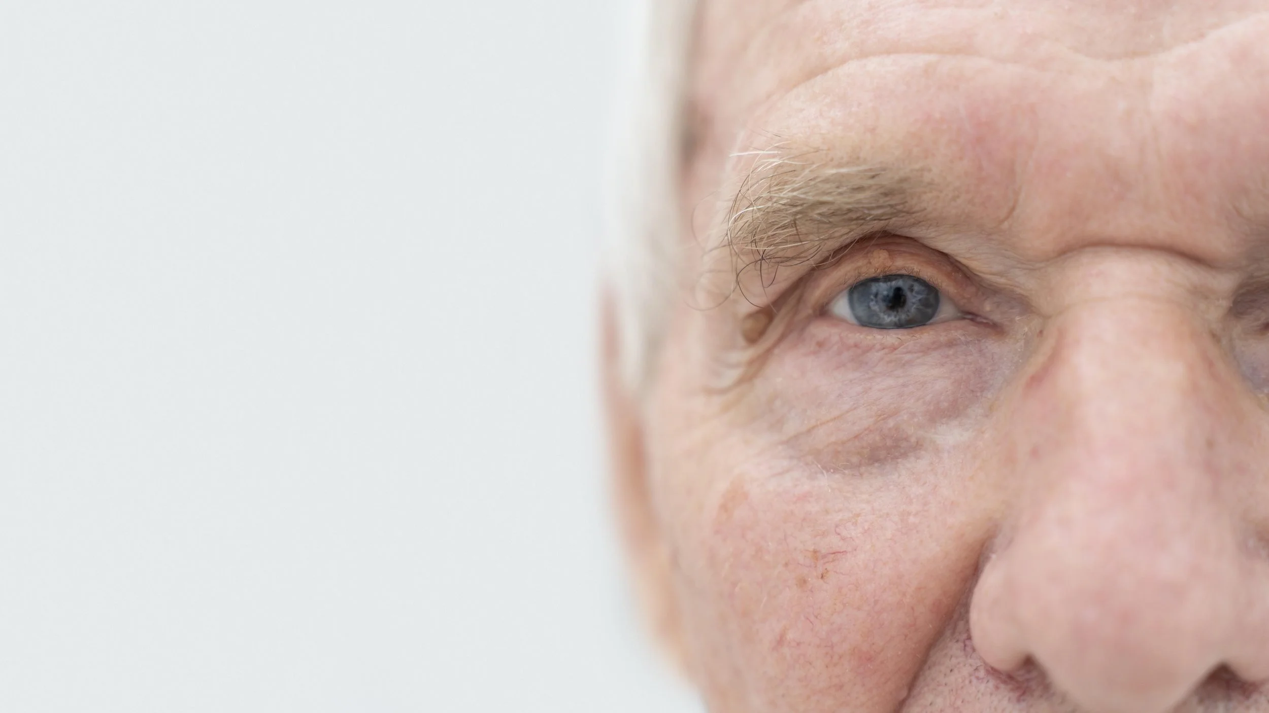 Close-up of an elderly man's face, showing blue eye, wrinkles, and light skin tone, against a plain light background.