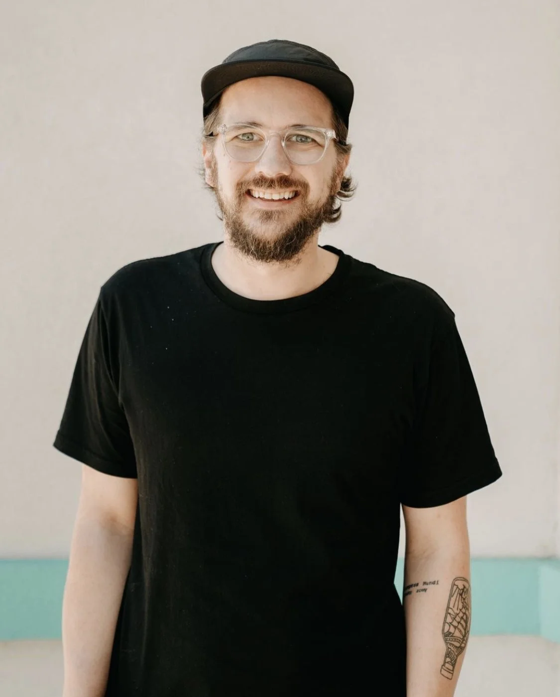 A young man with glasses, a beard, and tattoos on his arm, wearing a black hat and black t-shirt, smiling against a plain background.