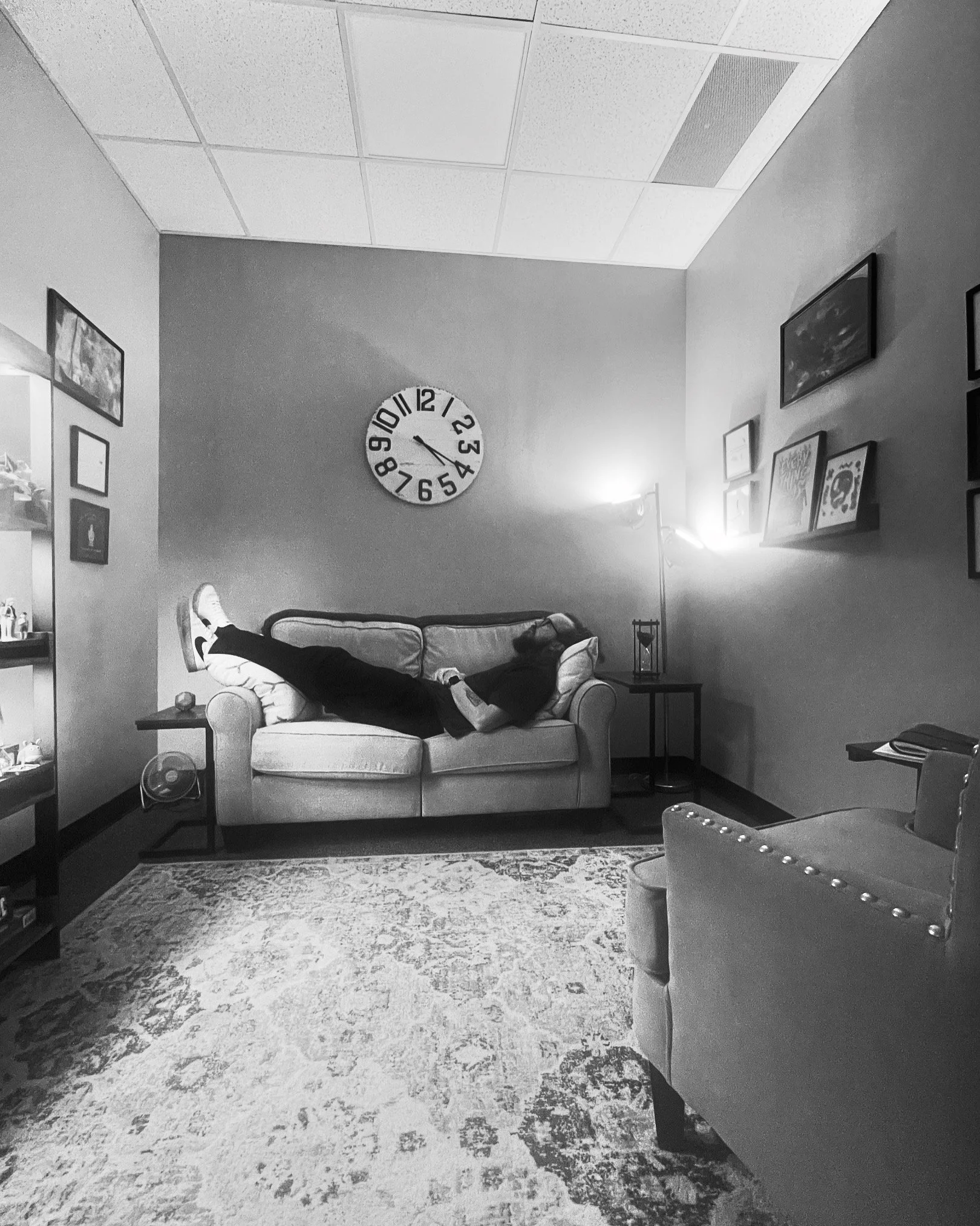 A living room with a couch, a wall clock, framed pictures, a lamp, and patterned carpet, with a person lying on the couch.