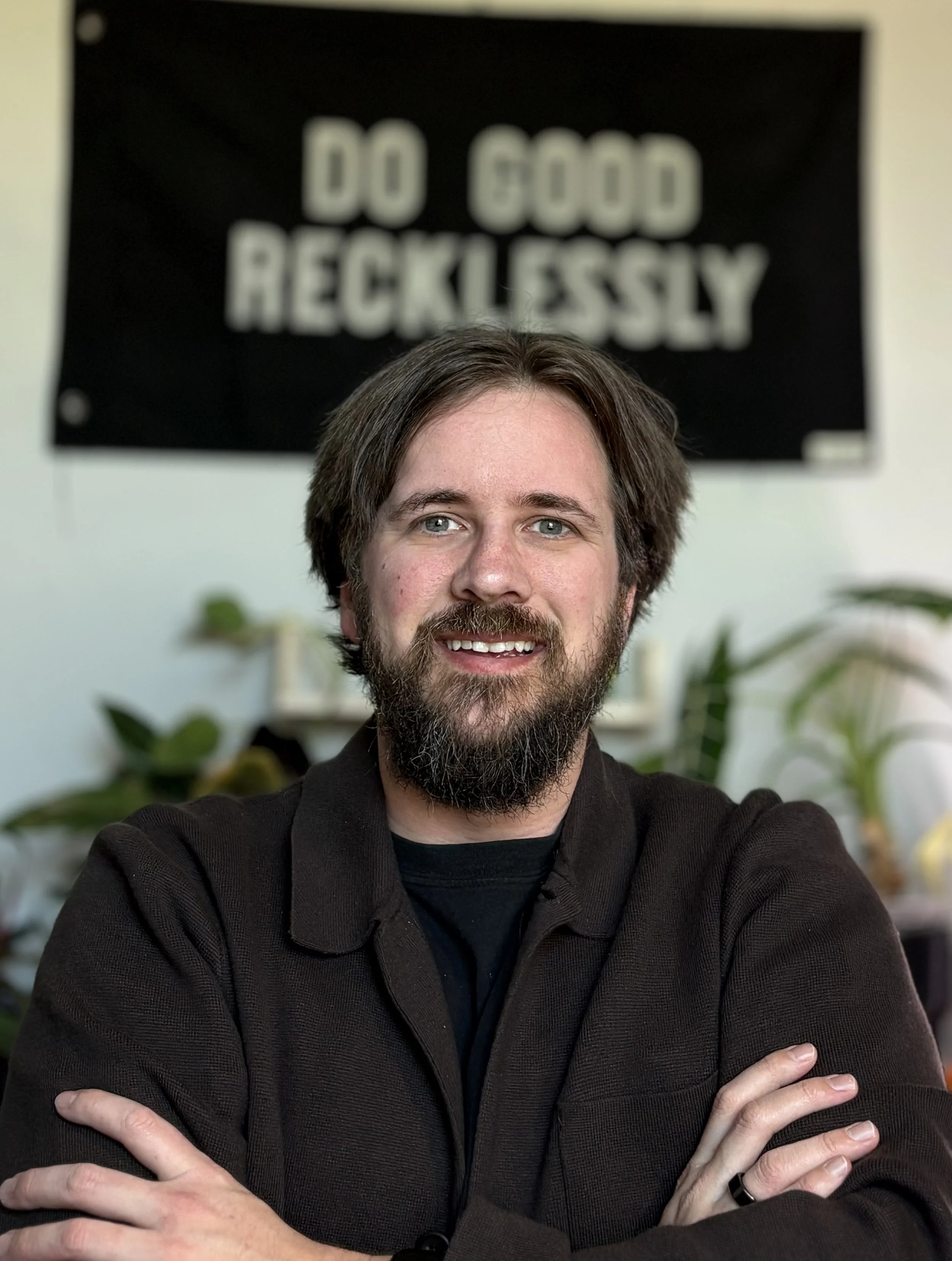 A man with brown hair, blue eyes, and a beard is smiling and has his arms crossed. He is wearing a black shirt and standing in front of a wall with a sign that says "DO GOOD RECKLESSLY".