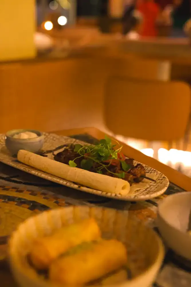 A plate with grilled skewers topped with green herbs, a breadstick, and a small bowl of sauce or dip on a restaurant table. In the foreground, a bowl with fried items, likely plantains or chips.