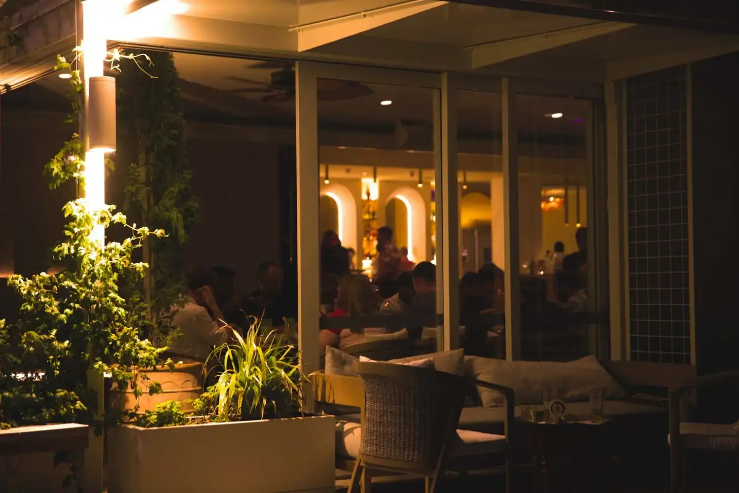 Night view of a restaurant patio with potted plants and seating, visible through glass walls with warm lighting inside, and people dining.