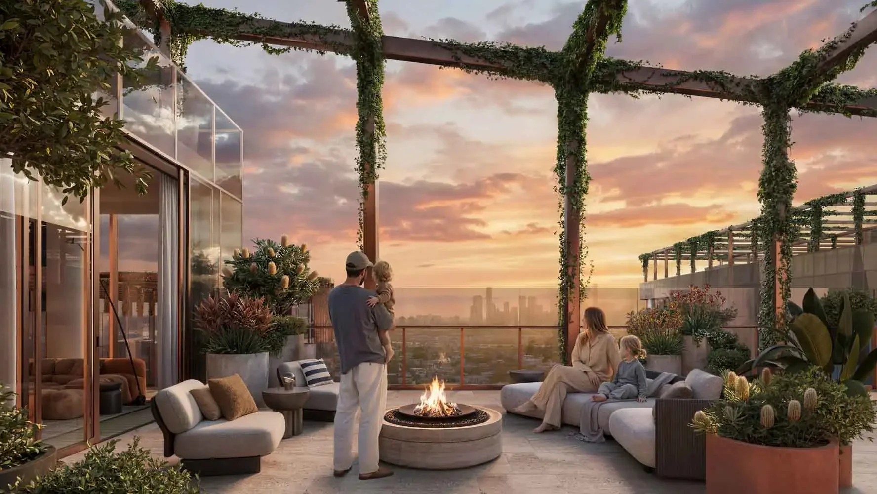 A rooftop patio with a fire pit, surrounded by comfortable seating, and decorated with numerous plants. Four people are present: a man holding a child, and a woman with a young girl sitting on a sofa. The sky is orange and pink at sunset, and the city skyline is visible in the distance.