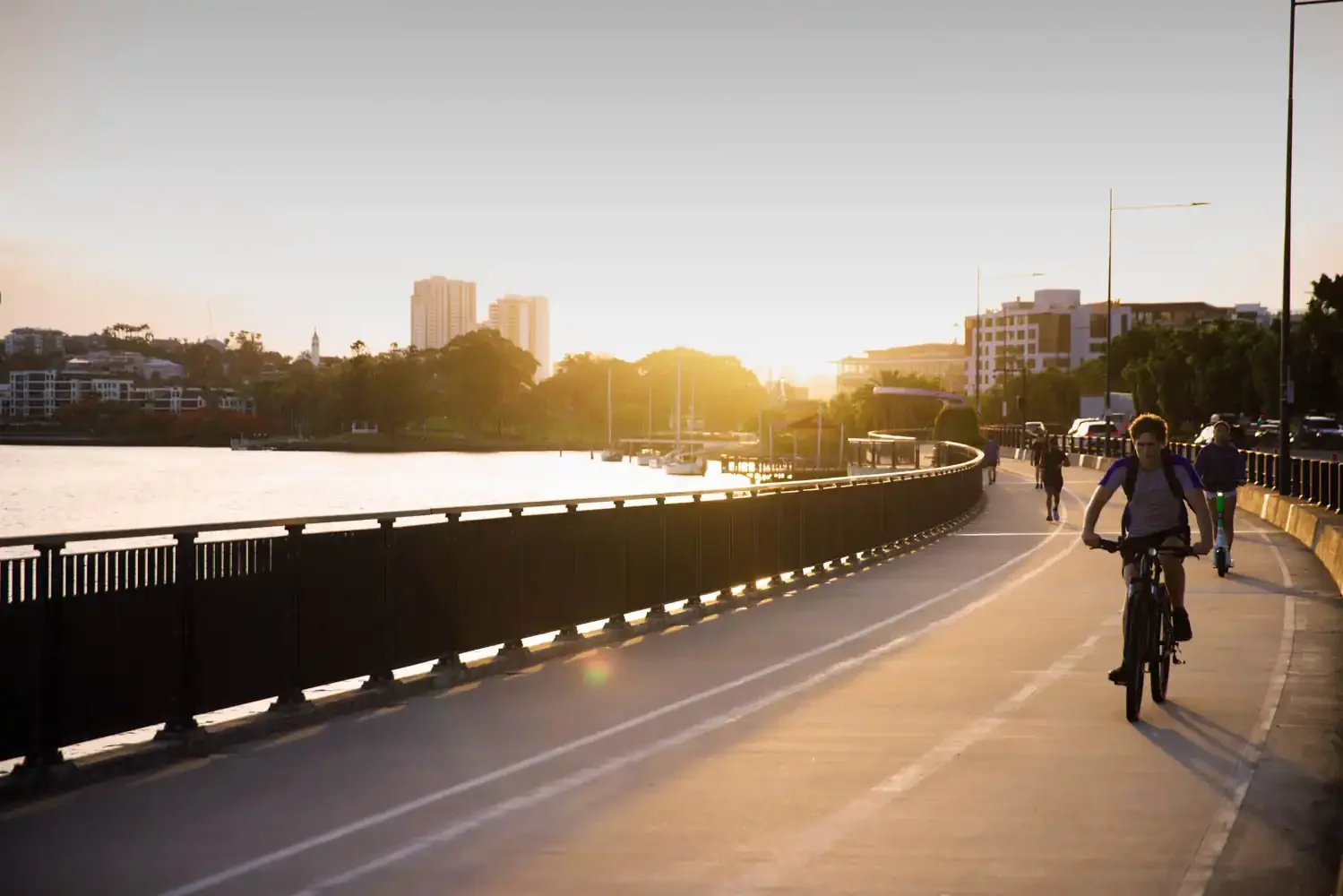 People riding bikes along a riverside pathway during sunset, with city buildings and trees in the background.