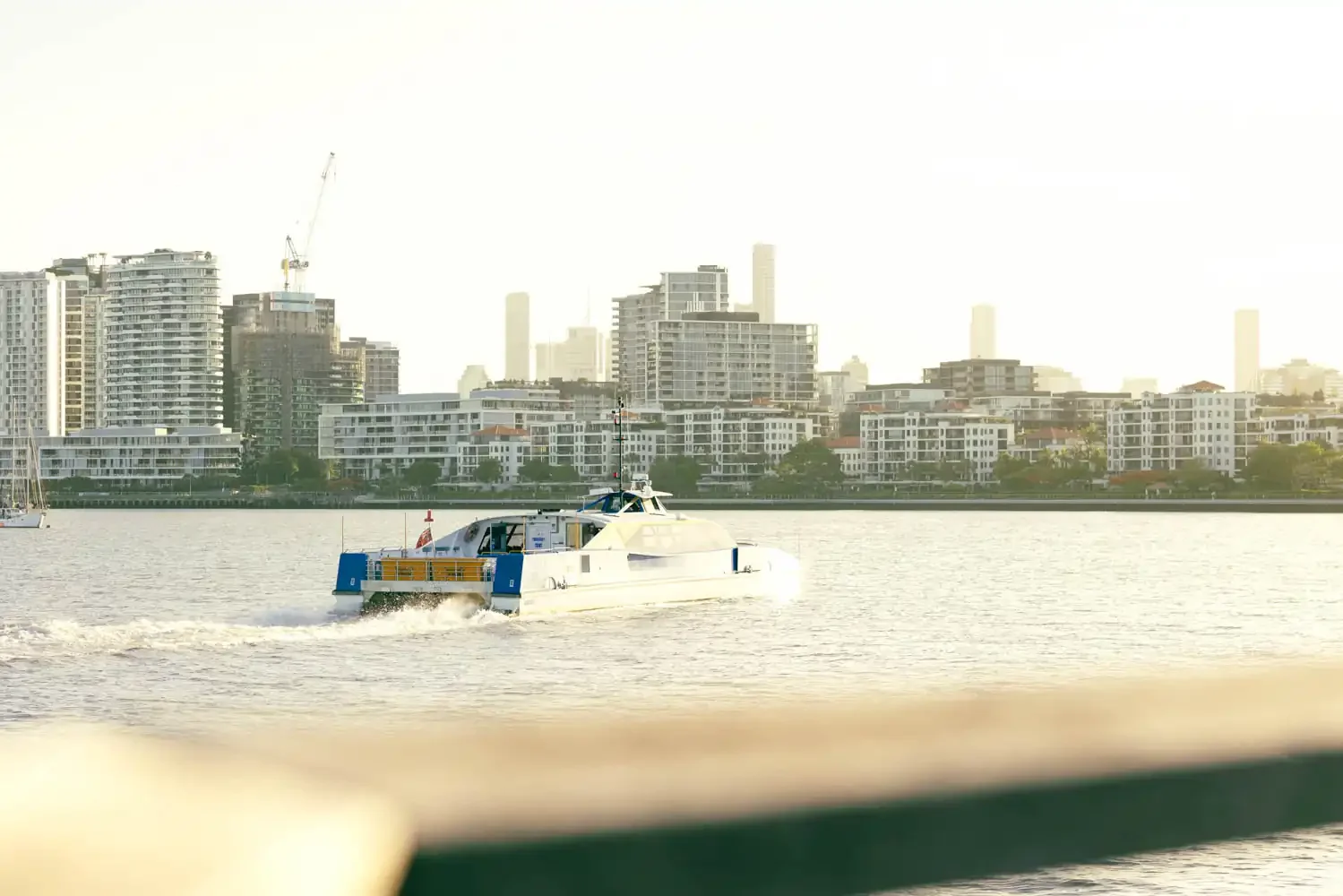 A white and blue boat moving across a body of water with a city skyline of high-rise buildings in the background.