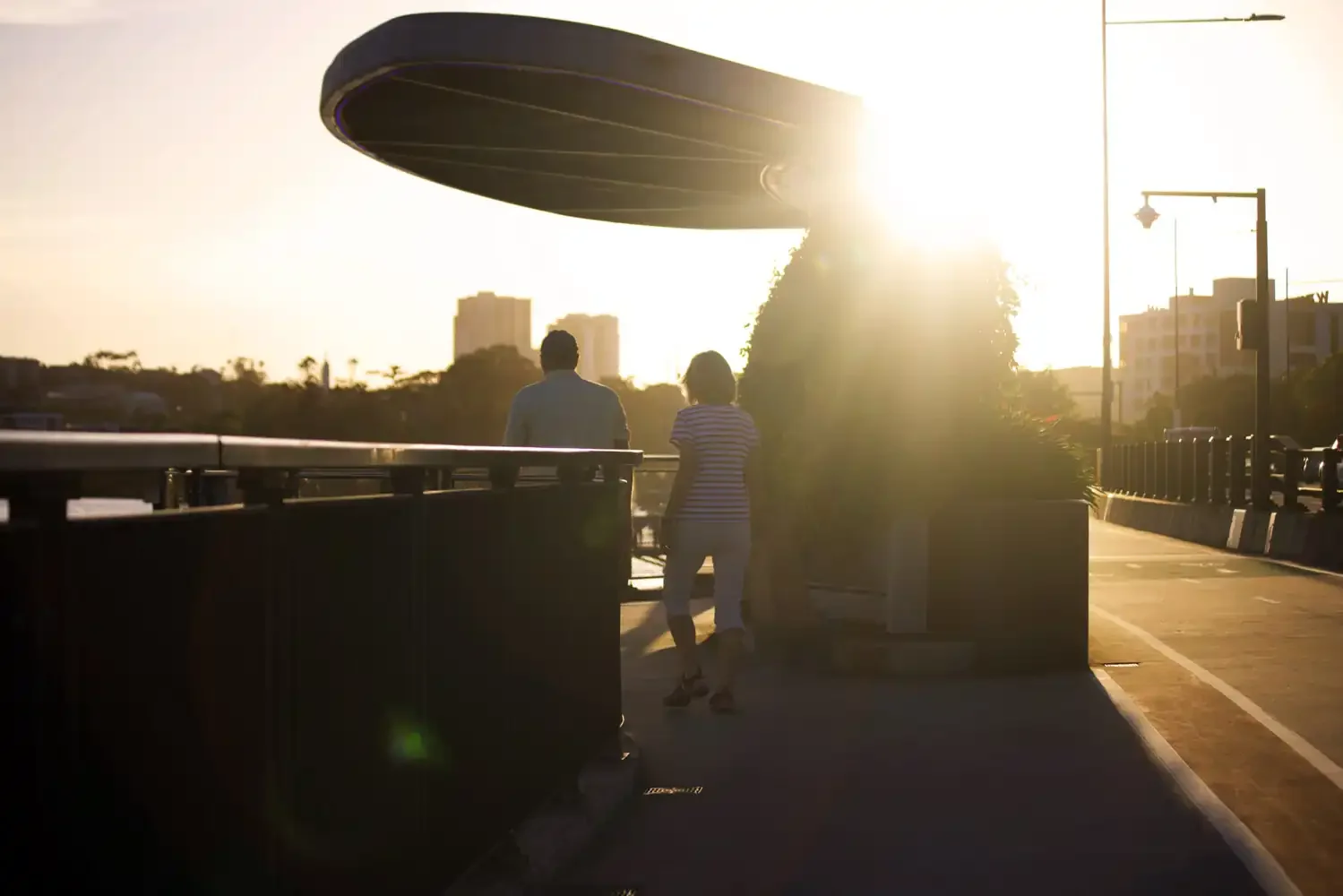 Two people standing on a sidewalk near a bus stop, with the sun setting in the background and a large curved building overhead.