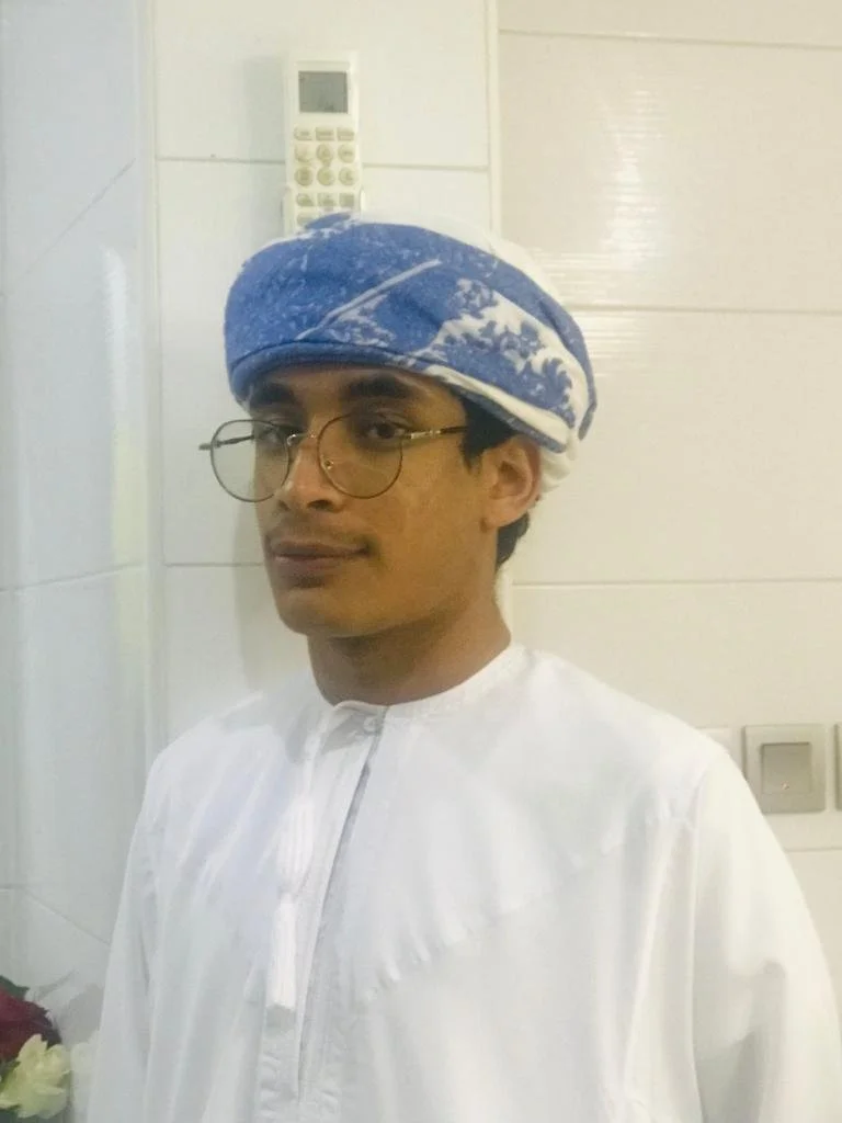 A man wearing traditional white Omani attire, glasses, and a blue and white patterned turban standing indoors in front of a white tiled wall.